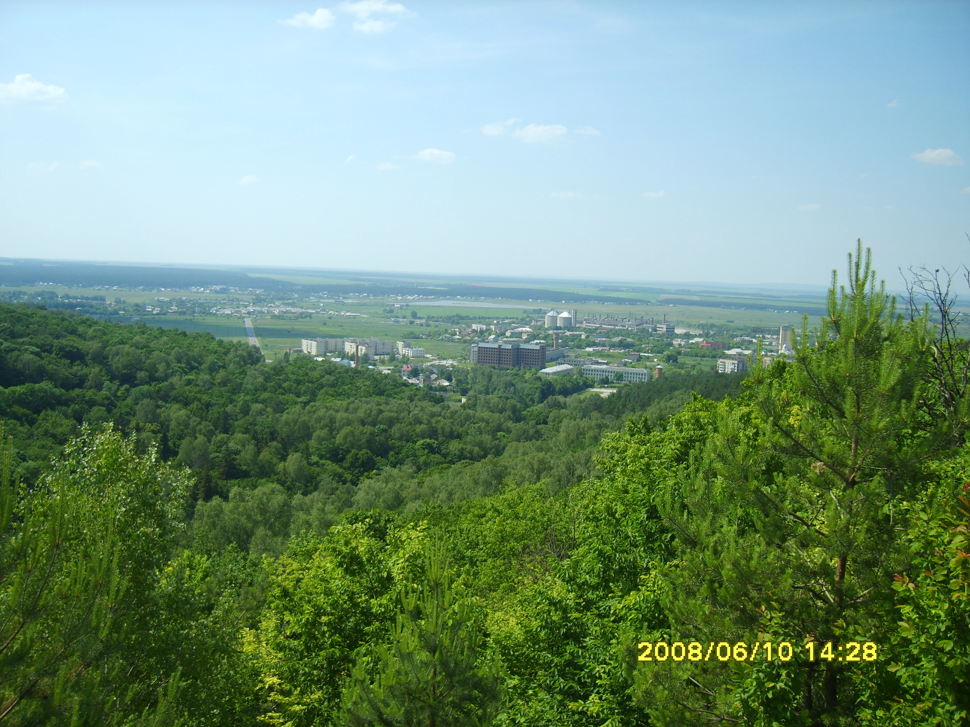 Lush green forested hills overlooking a valley with distant buildings under a clear blue sky