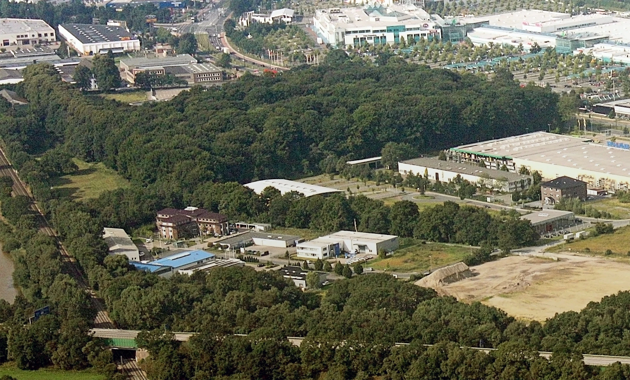 An aerial view showing a dense patch of forest surrounded by industrial buildings, warehouses, and transport infrastructure.