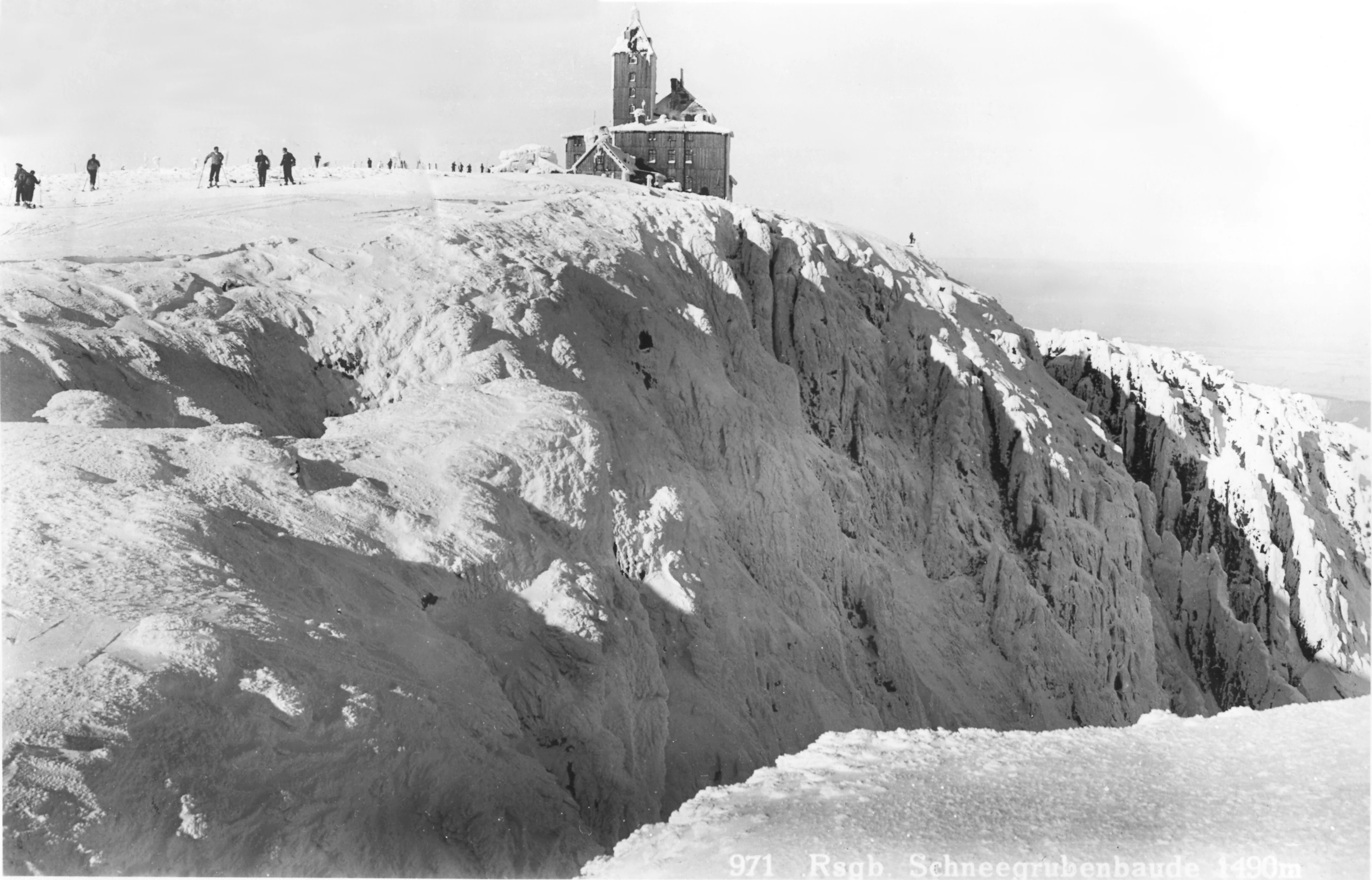 Black-and-white photograph of a snow-covered mountain cliff with a small building on the ridge, several people walking along the crest, and a steep drop into a valley below.