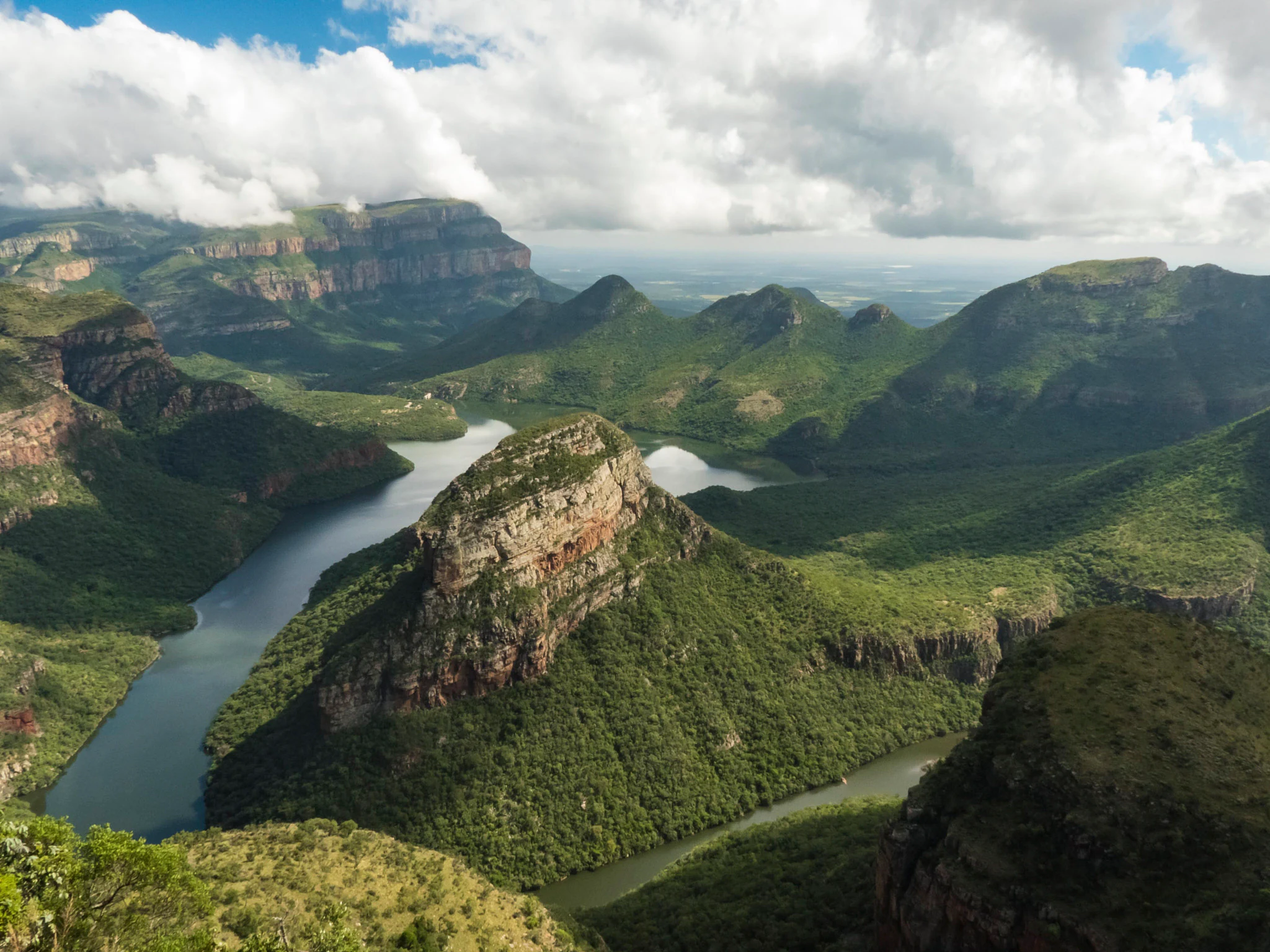 A wide aerial view looking down into the lush green Blyde River Canyon, with a river winding through the steep rocky valley and clouds resting on the distant mountain peaks.