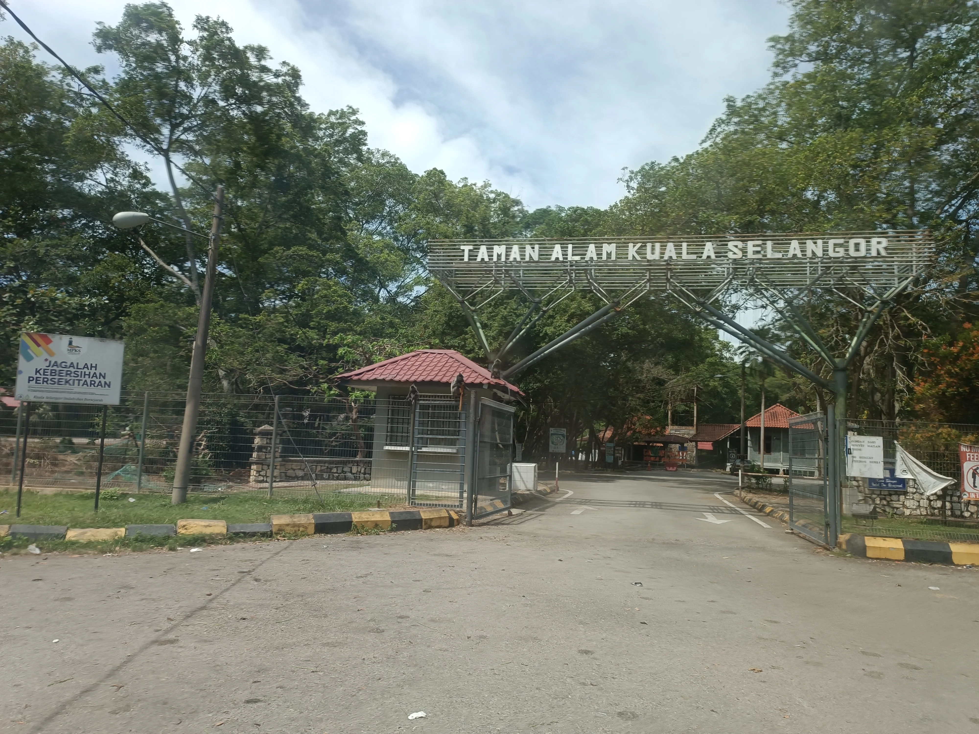 A paved road leads through a metal gate archway labeled Taman Alam Kuala Selangor, flanked by trees and a small guardhouse building.