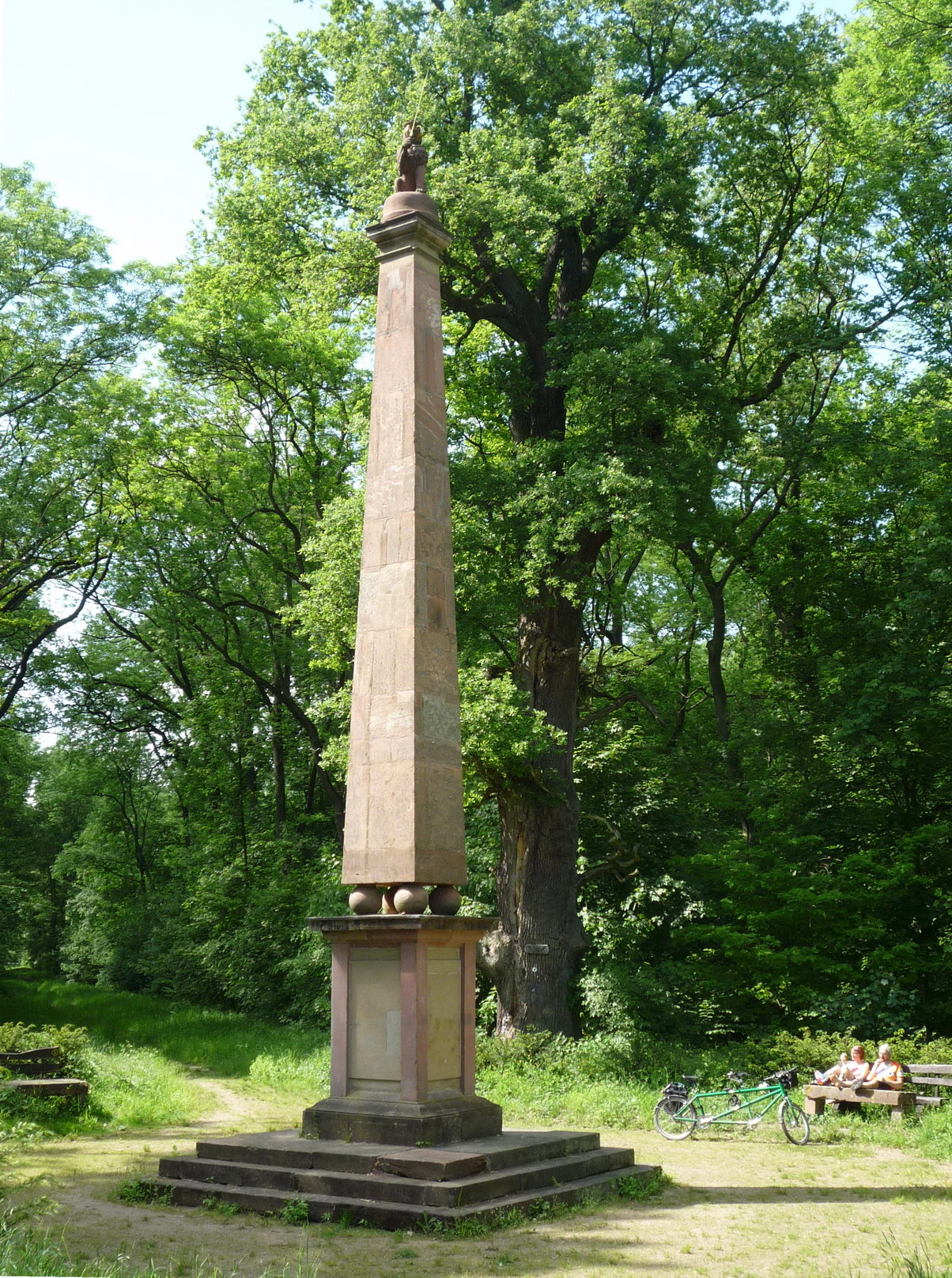 A tall, stone obelisk monument stands in a clearing surrounded by lush green trees, with a bicycle and people sitting on a bench visible nearby.
