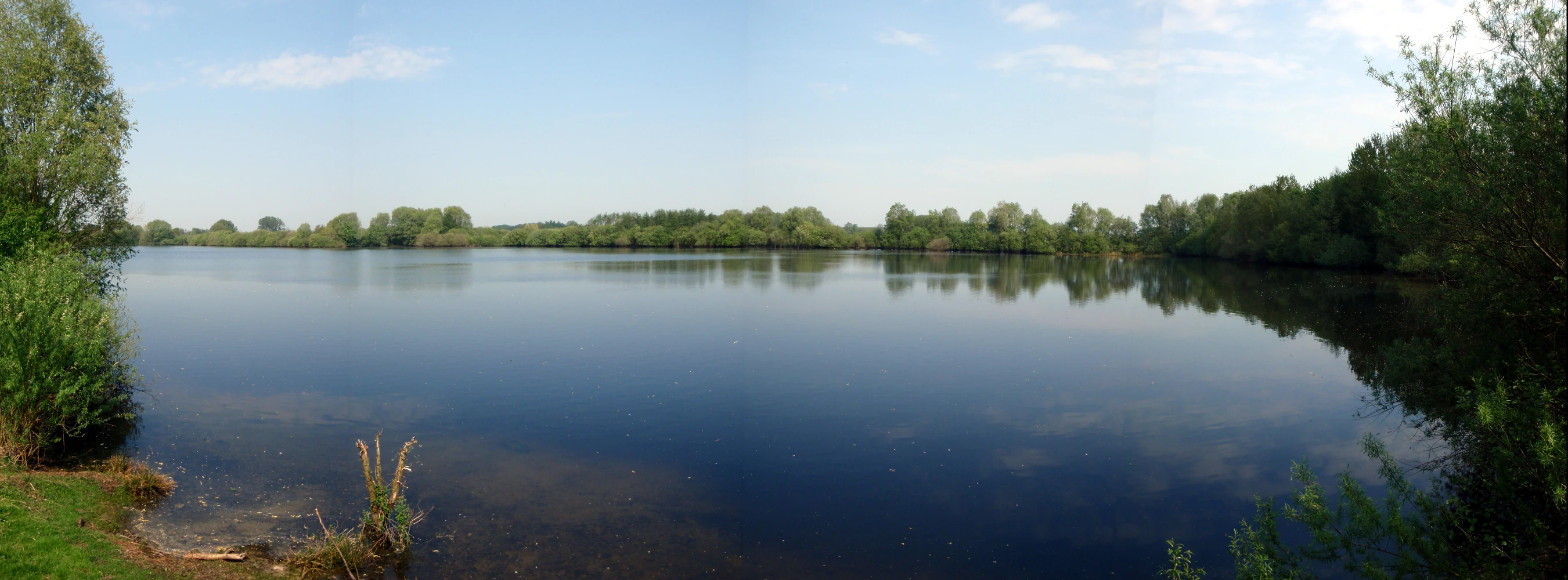 Una amplia toma panorámica de un lago tranquilo rodeado de árboles densos y arbustos bajo un cielo azul despejado en Kuhlenvenn.