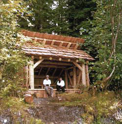 Dos personas se sientan en un banco de madera dentro de un pequeño refugio rústico de madera en el bosque, con techo de tejas, rodeado de árboles.