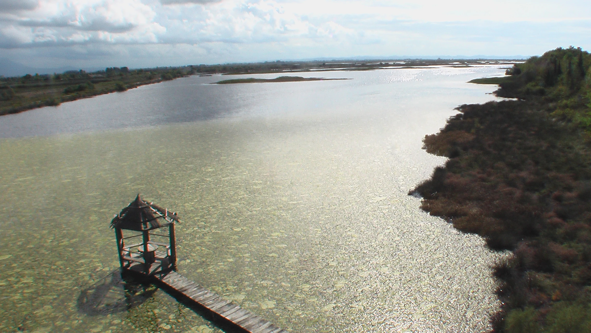 A high-angle view of a calm lagoon with a wooden pier and a small covered observation structure extending into the water, bordered by dense vegetation.