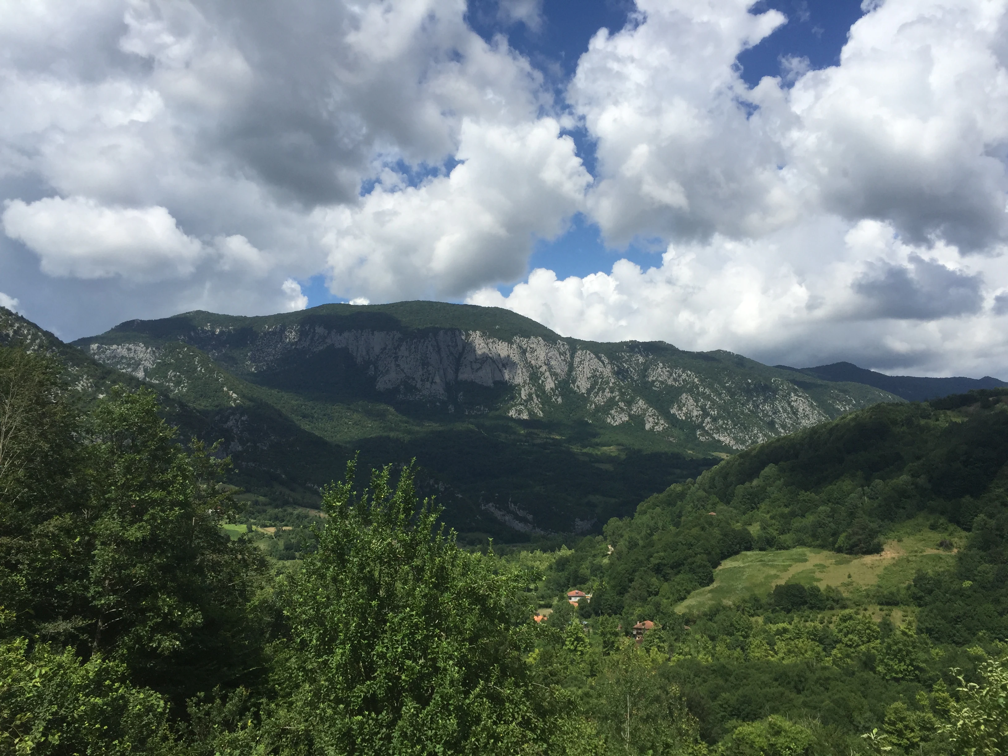 Mountainous landscape with green valleys, scattered trees, and a village in the distance under a partly cloudy sky