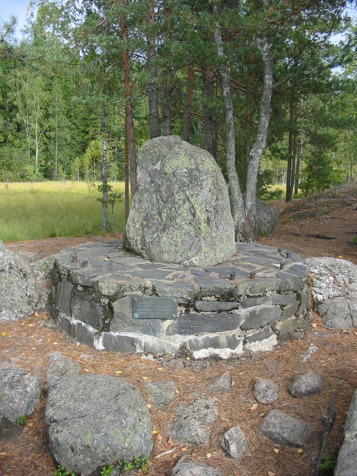 Large stone monument with a plaque surrounded by rocks and trees in a forested area