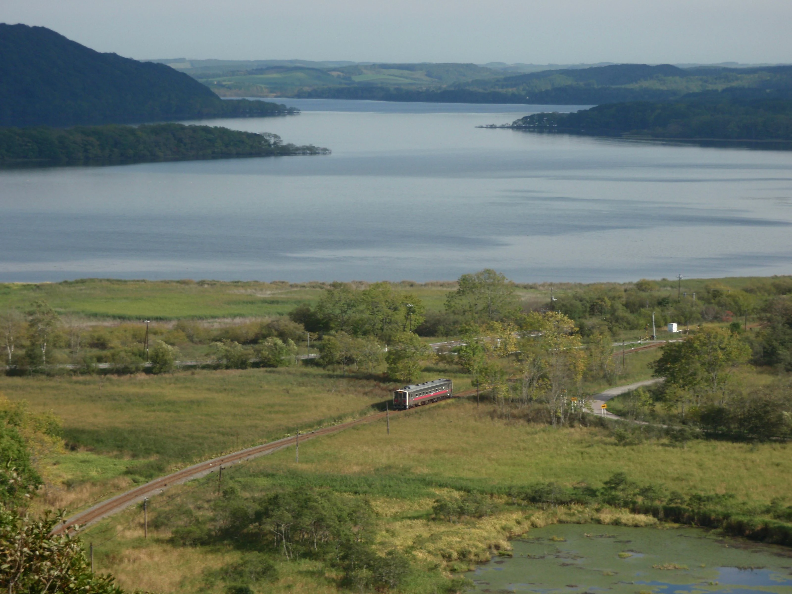 Una vista panorámica del lago Touro con una vía férrea que atraviesa humedales herbáceos, rodeado de colinas y vegetación