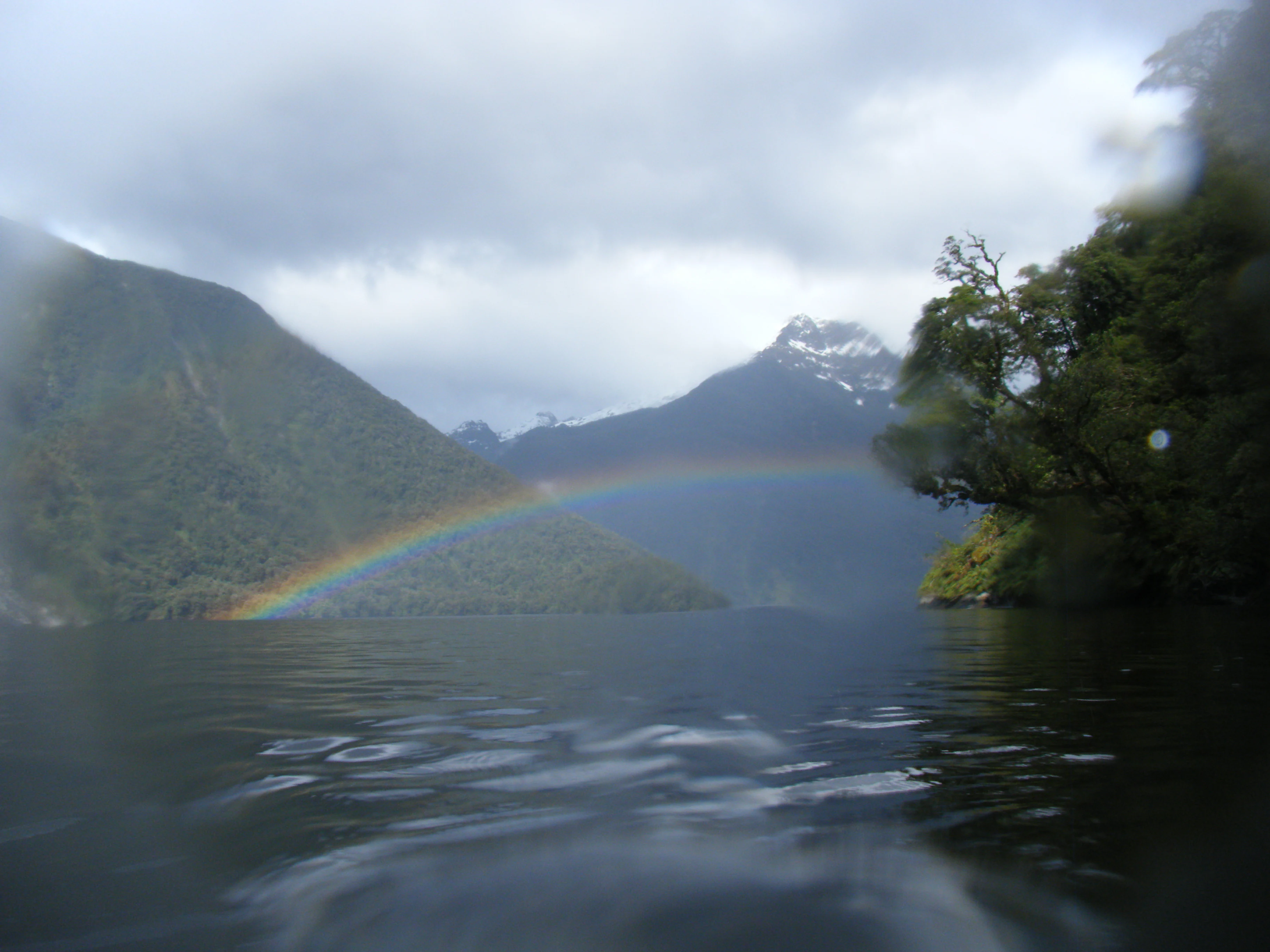 A low-angle view from the water looking toward a rainbow spanning across a quiet fiord, surrounded by steep, lush green, mountain slopes under a cloudy sky.