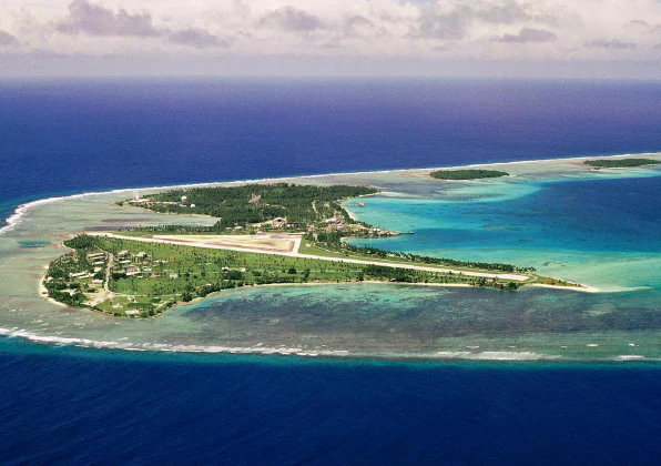 Una fotografía aérea de una isla coralina de baja altitud rodeada de aguas turquesa de laguna y océano azul profundo, con una pista de aterrizaje y estructuras dispersas.