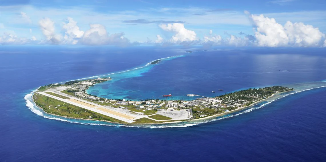 Una vista panorámica aérea de una isla coralina larga y estrecha rodeada de océano azul profundo, con una pista de aterrizaje y edificios de apoyo visibles en la isla.