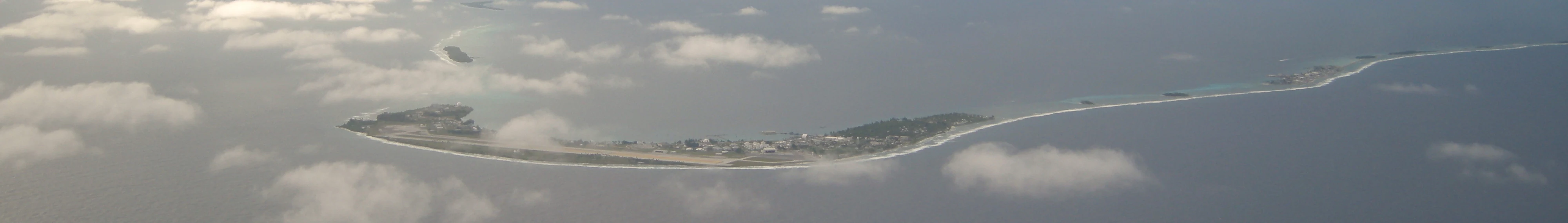 Vista aérea de un atolón de coral arenoso, delgado y curvo, rodeado de aguas oceánicas azul oscuro, parcialmente oculto por nubes blancas dispersas.