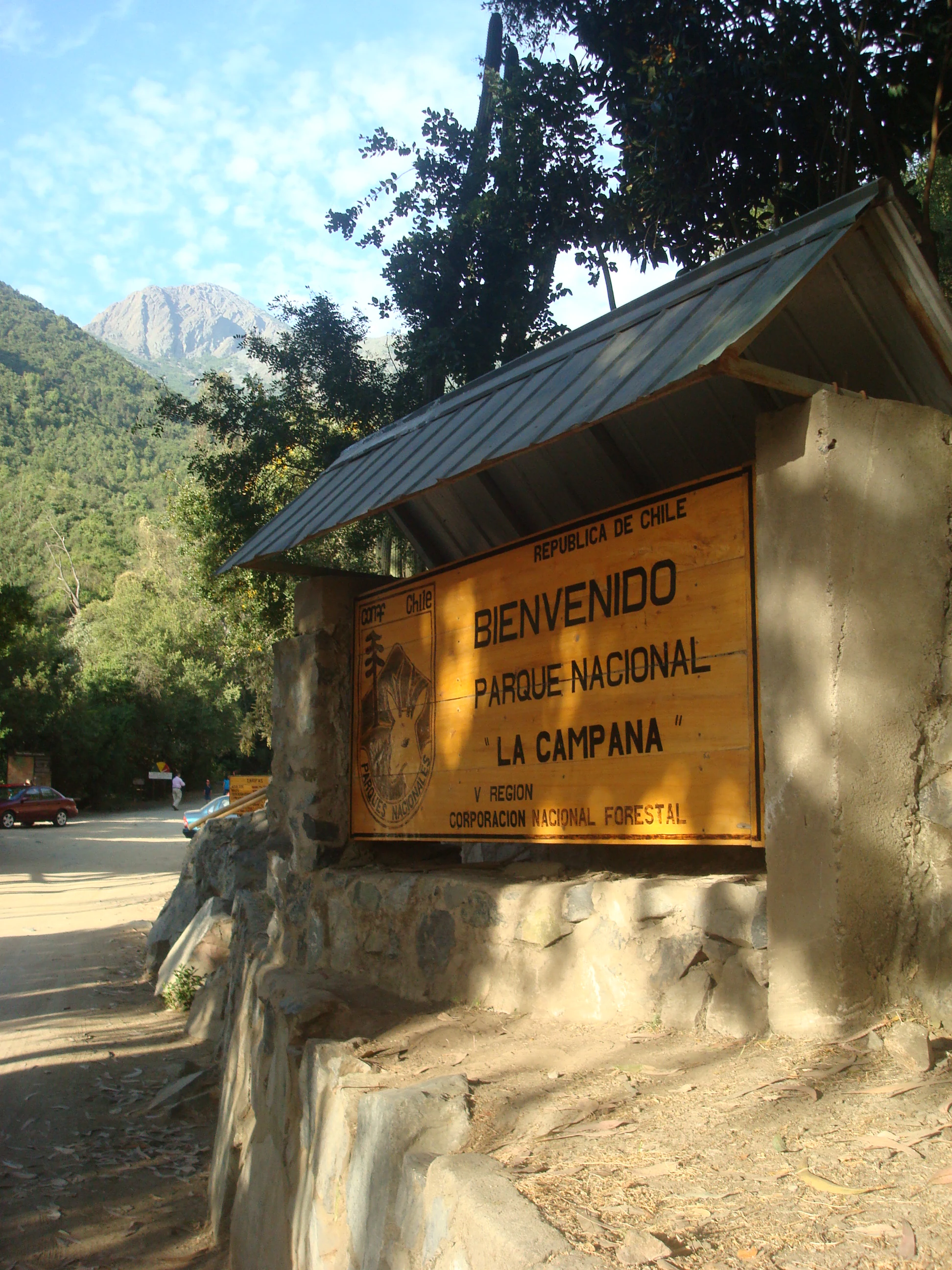 Yellow entrance sign for Parque Nacional La Campana under a metal roof, stone structure, with trees and mountain in background