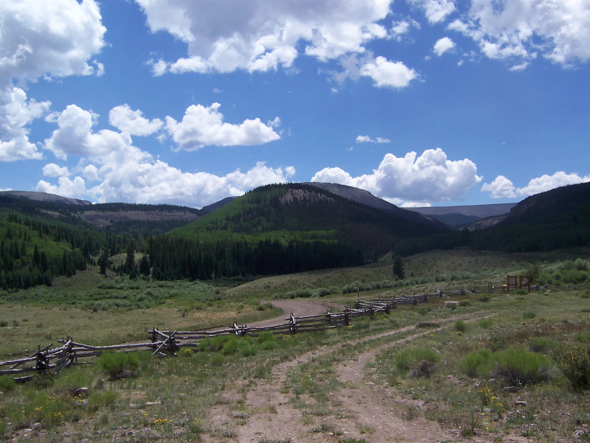 A wide landscape view of grassy plains leading to rolling mountains under a bright blue sky with puffy white clouds, featuring a rustic wooden fence in the foreground.