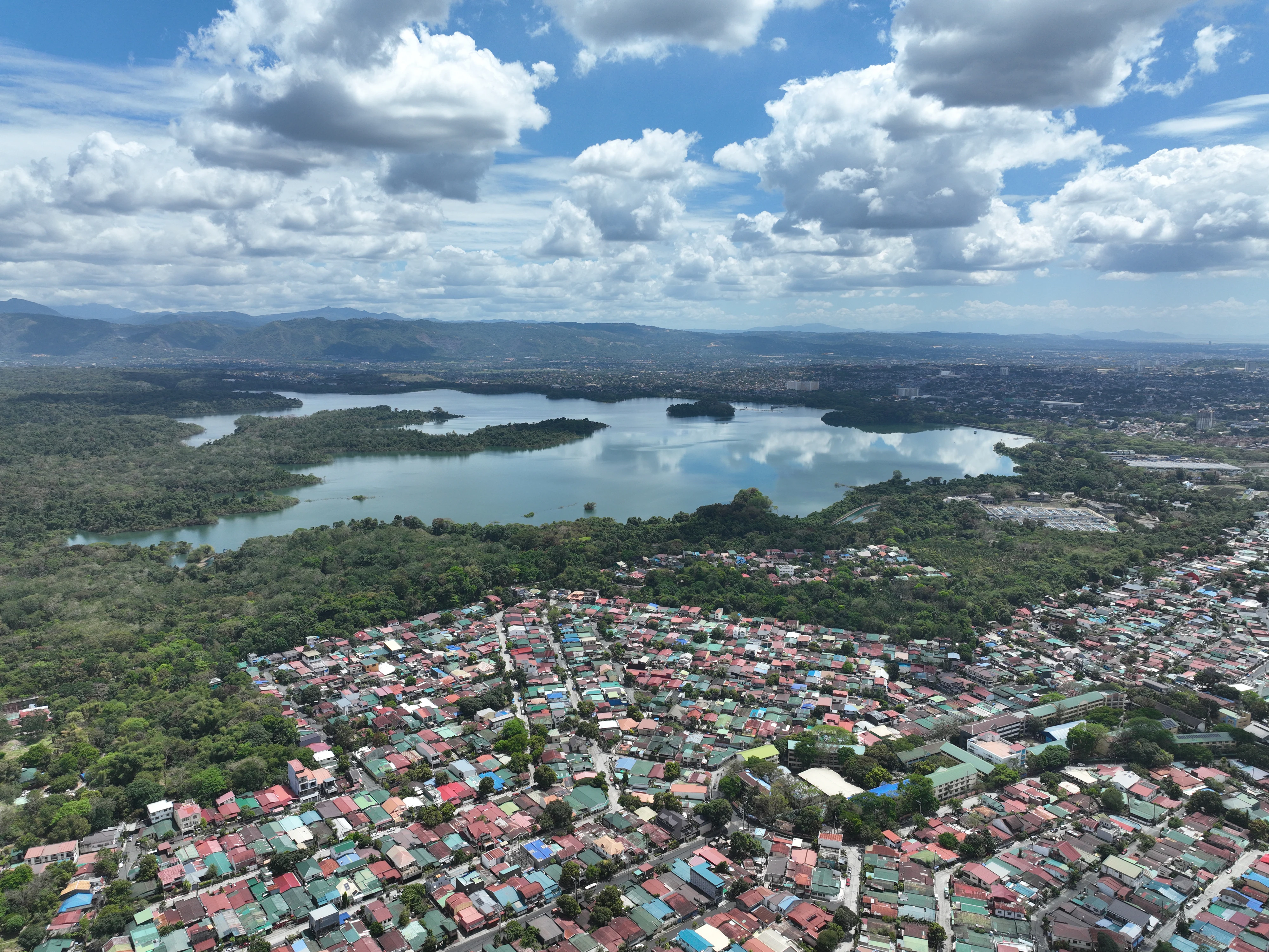 Aerial drone shot looking down at the large body of the La Mesa Dam reservoir surrounded by dense forest, bordered by a dense residential neighborhood in the foreground under a cloudy blue sky.