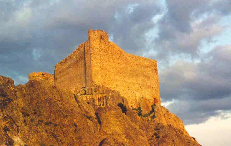 A medieval stone castle fortress sits on a craggy hilltop at sunset, illuminated by warm golden light against a cloudy sky.