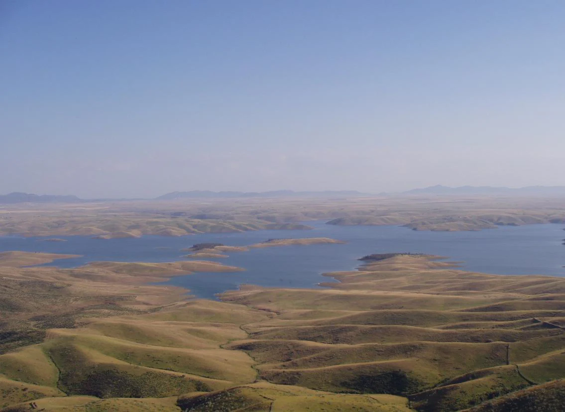 A wide, elevated landscape view featuring a large blue reservoir winding through rolling, dry golden hills under a clear sky.