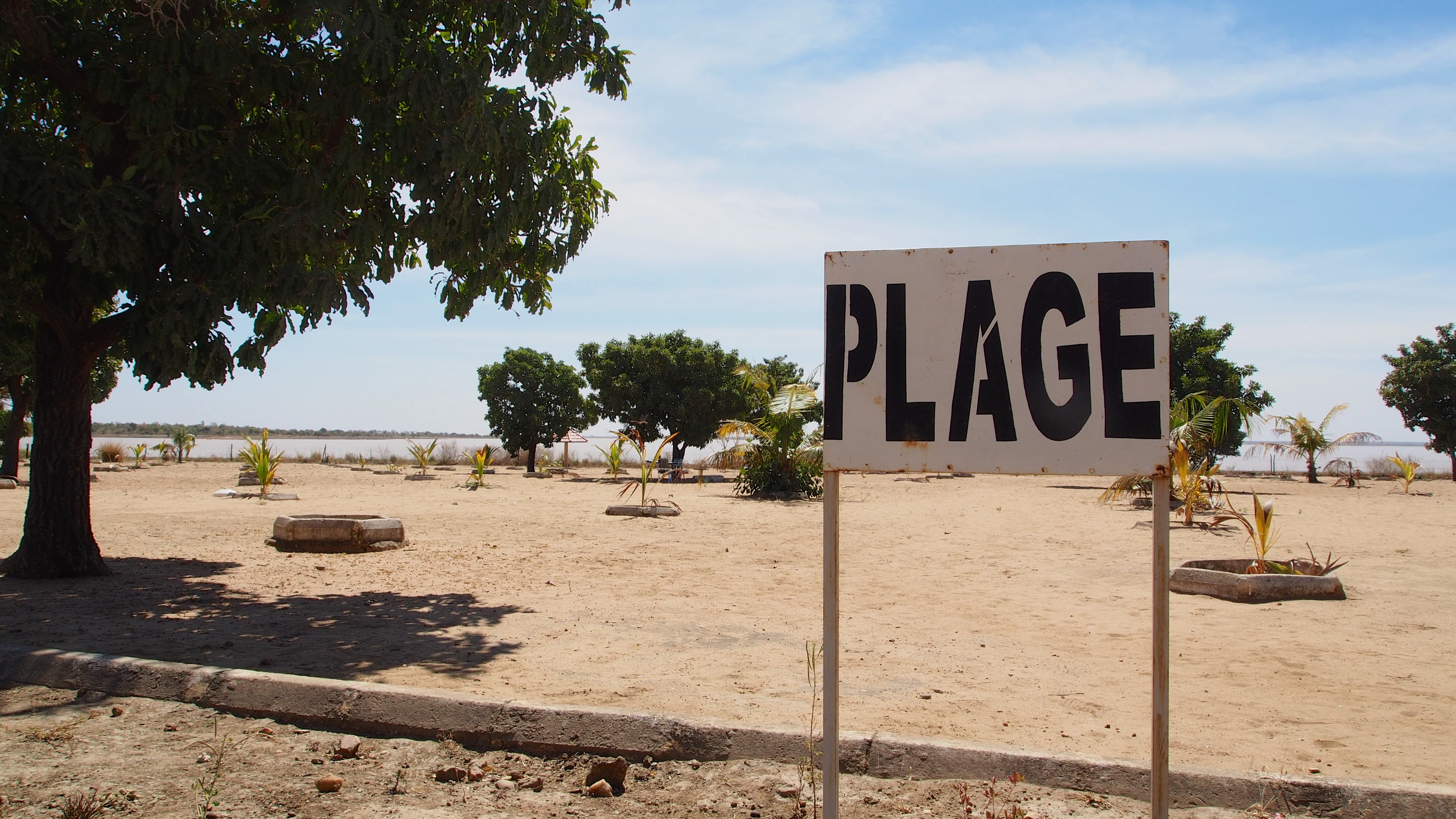 Un cartel blanco con grandes letras negras que dicen PLAGE situado en una zona arenosa cerca de árboles y un lago.