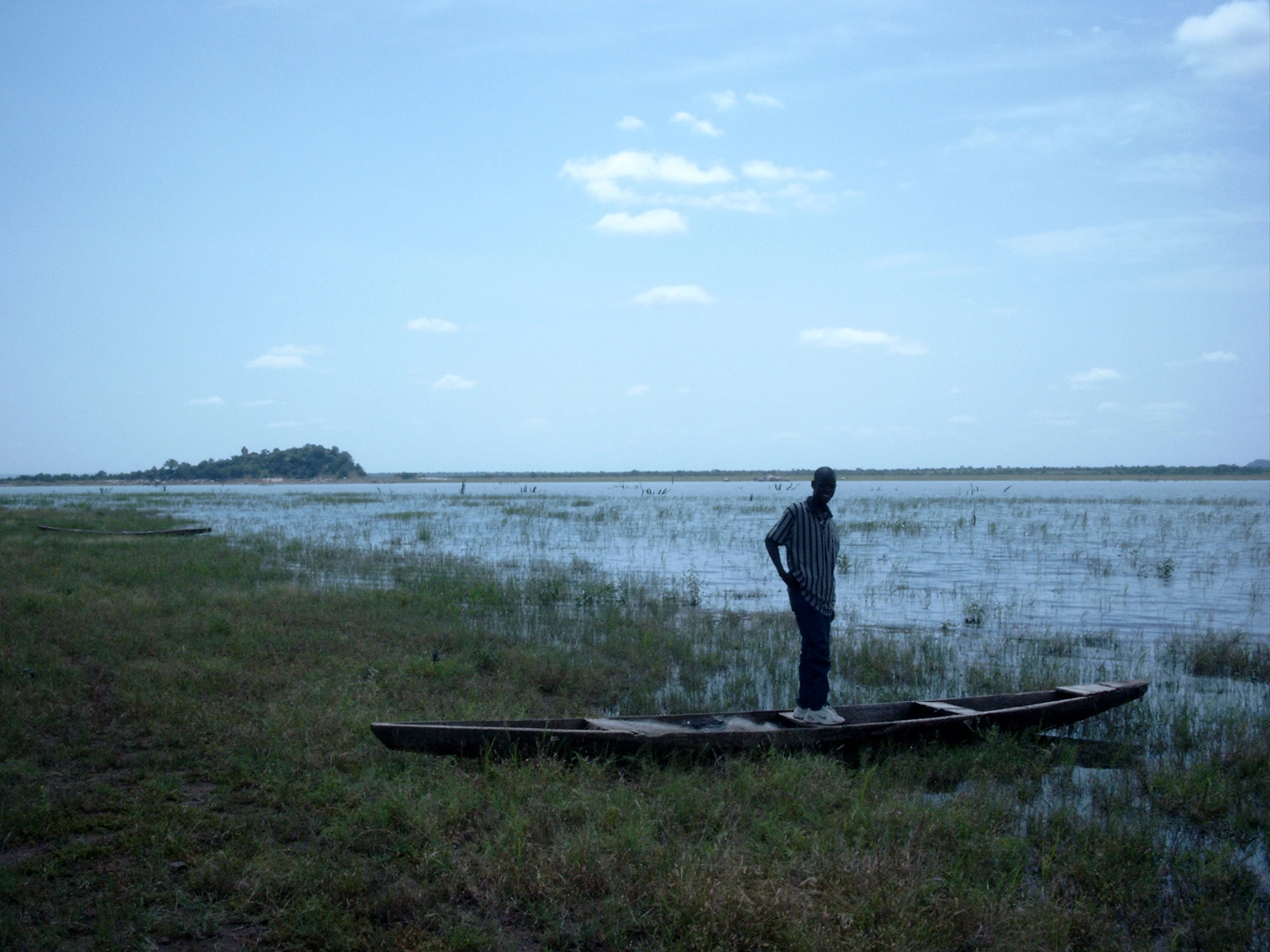 A person stands in a shallow, narrow wooden boat pulled up onto the grassy, marshy shoreline of a large lake under a pale blue sky.