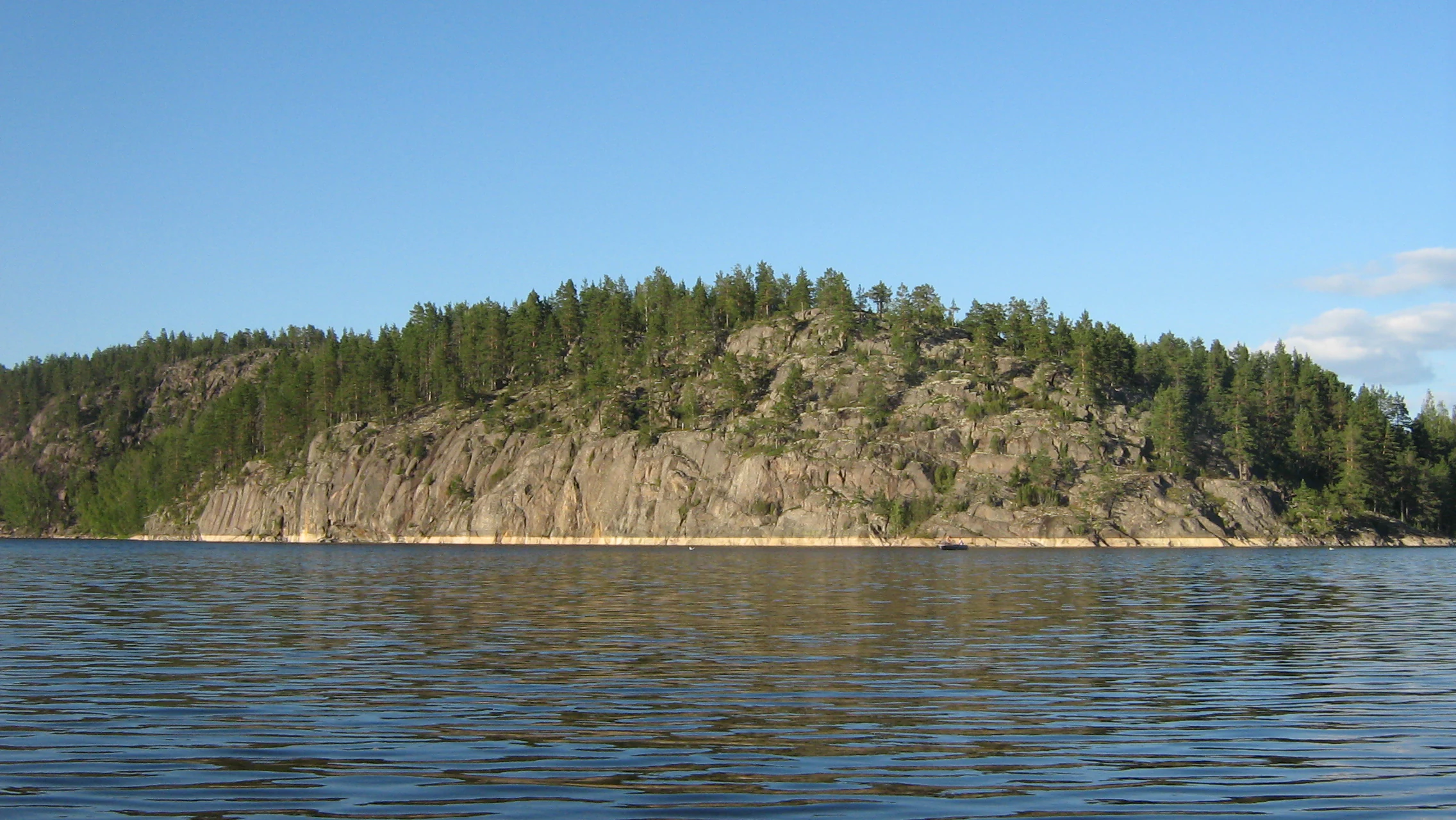 A low, rocky island covered in pine forest rises from the surface of calm water under a clear blue sky, illustrating the skerry geography of Lake Ladoga.