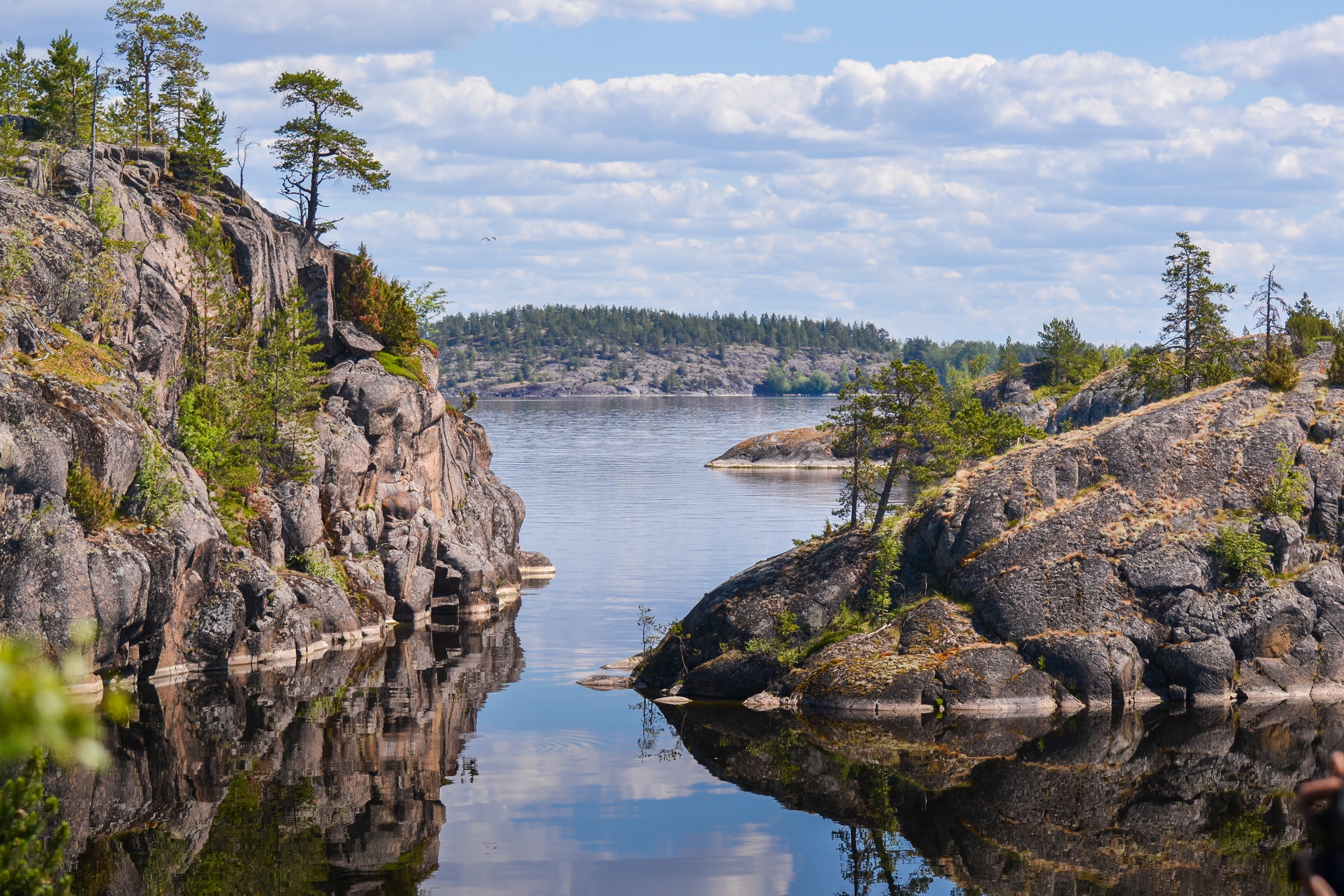 A scenic landscape view of the Ladozhskiye Shkhery National Park showing rocky, pine-covered cliffs bordering calm, reflective water under a sky with scattered clouds.
