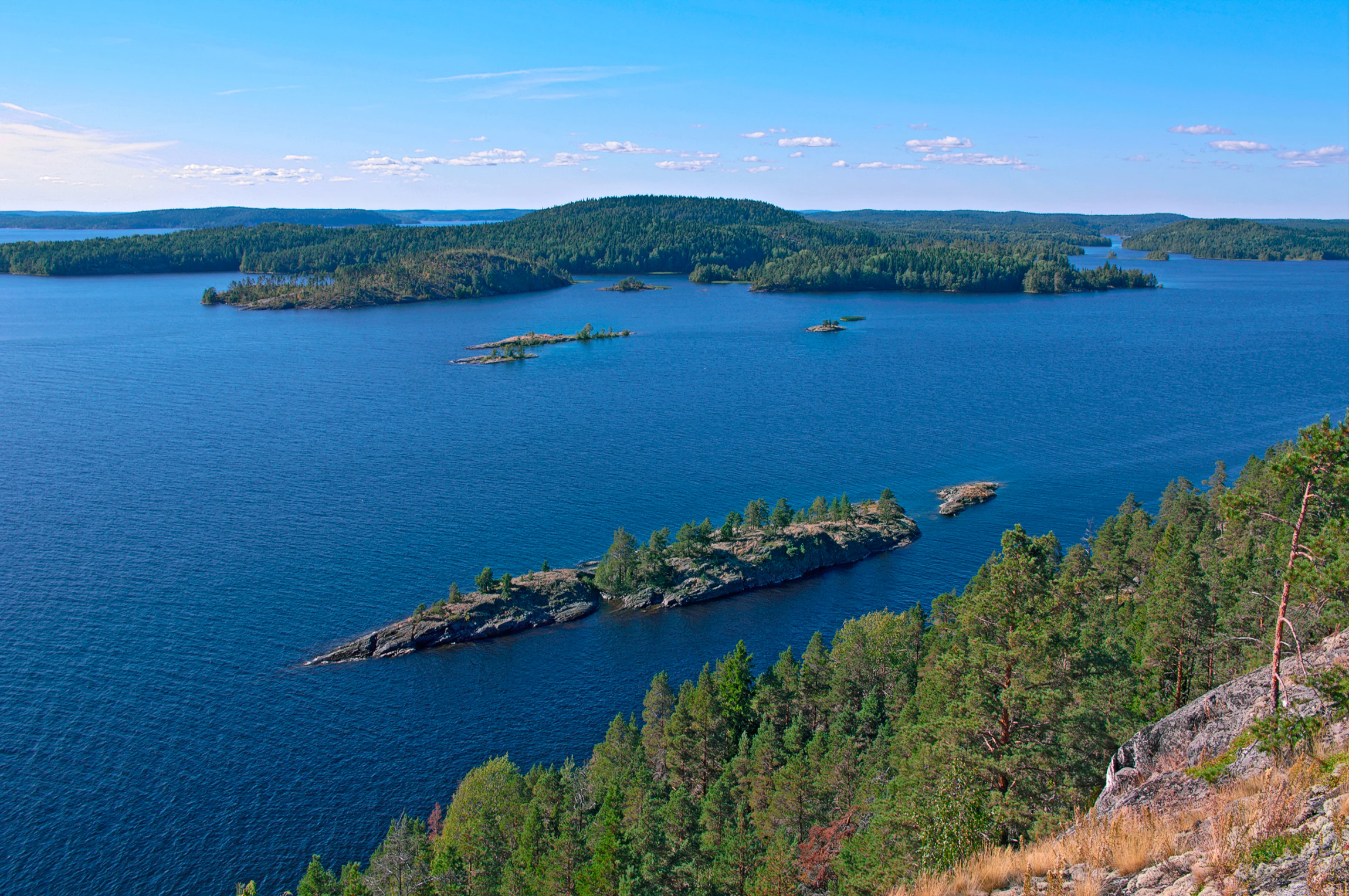A high angle panoramic view overlooking calm blue water dotted with multiple small rocky islands covered in green pine forests under a clear blue sky.