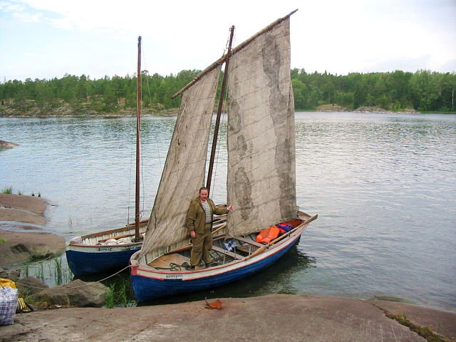 A person stands on a blue, traditional wooden boat with sails moored against a rocky shoreline on a lake, with a second similar boat visible behind it.