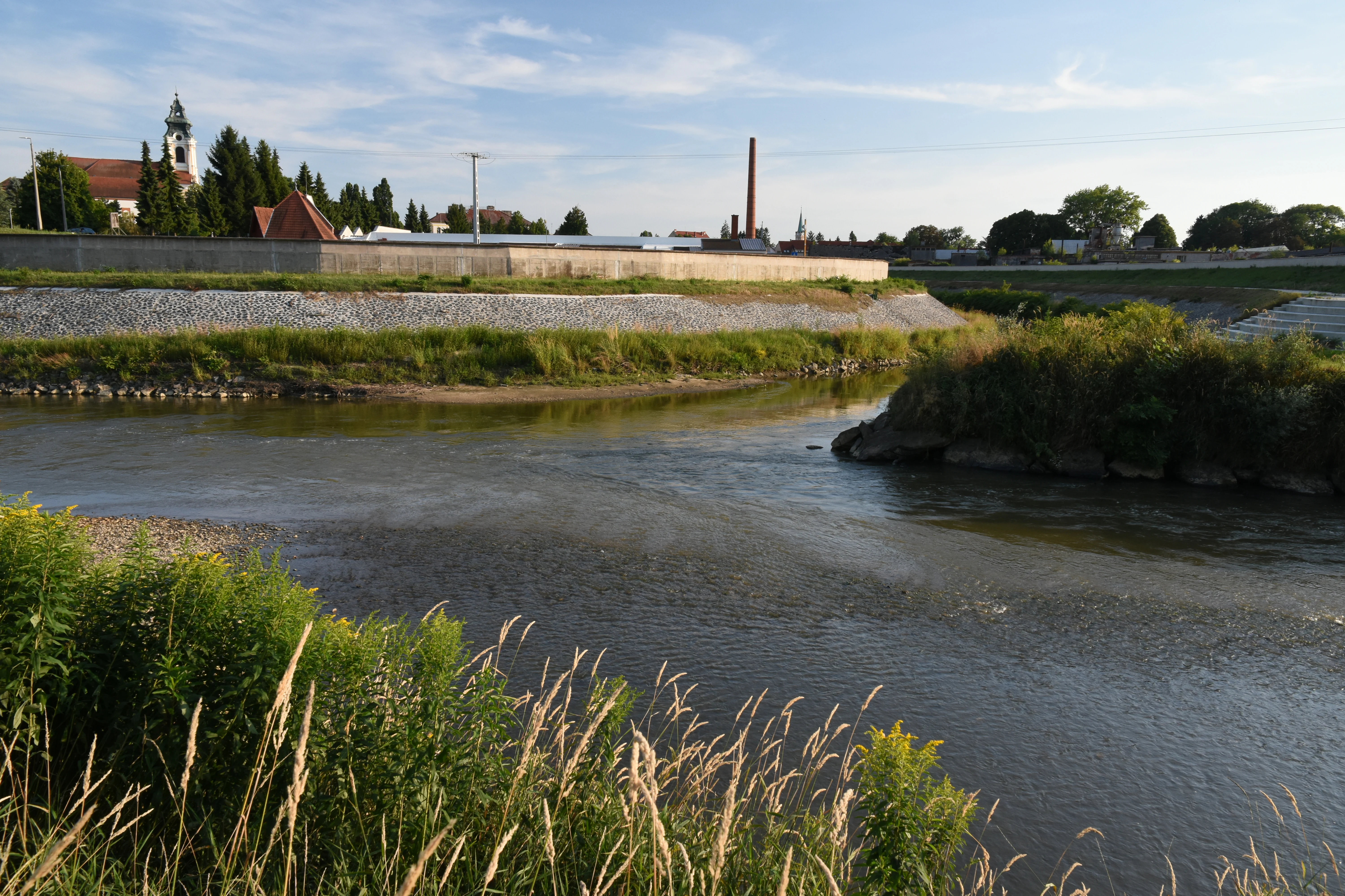 A river confluence where two waterways join, featuring a grassy embankment and a church tower visible in the background.