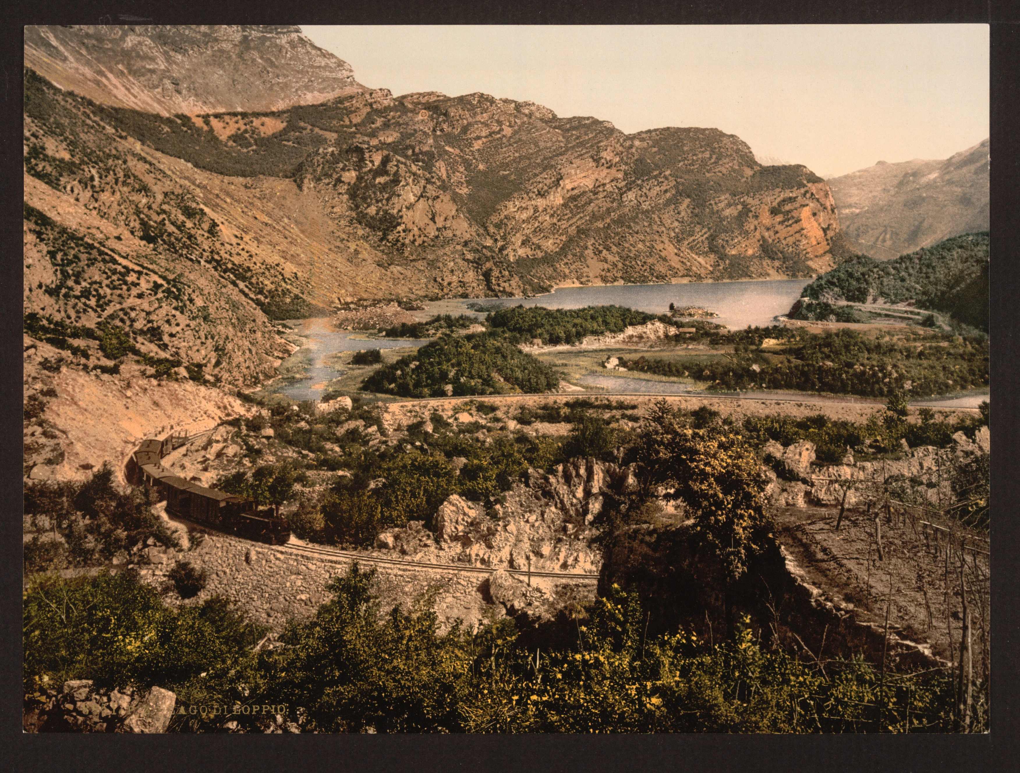 A vintage photographic print showing a valley with a lake, marshy vegetation, and rocky mountains in the background, with a train visible on a track in the foreground.