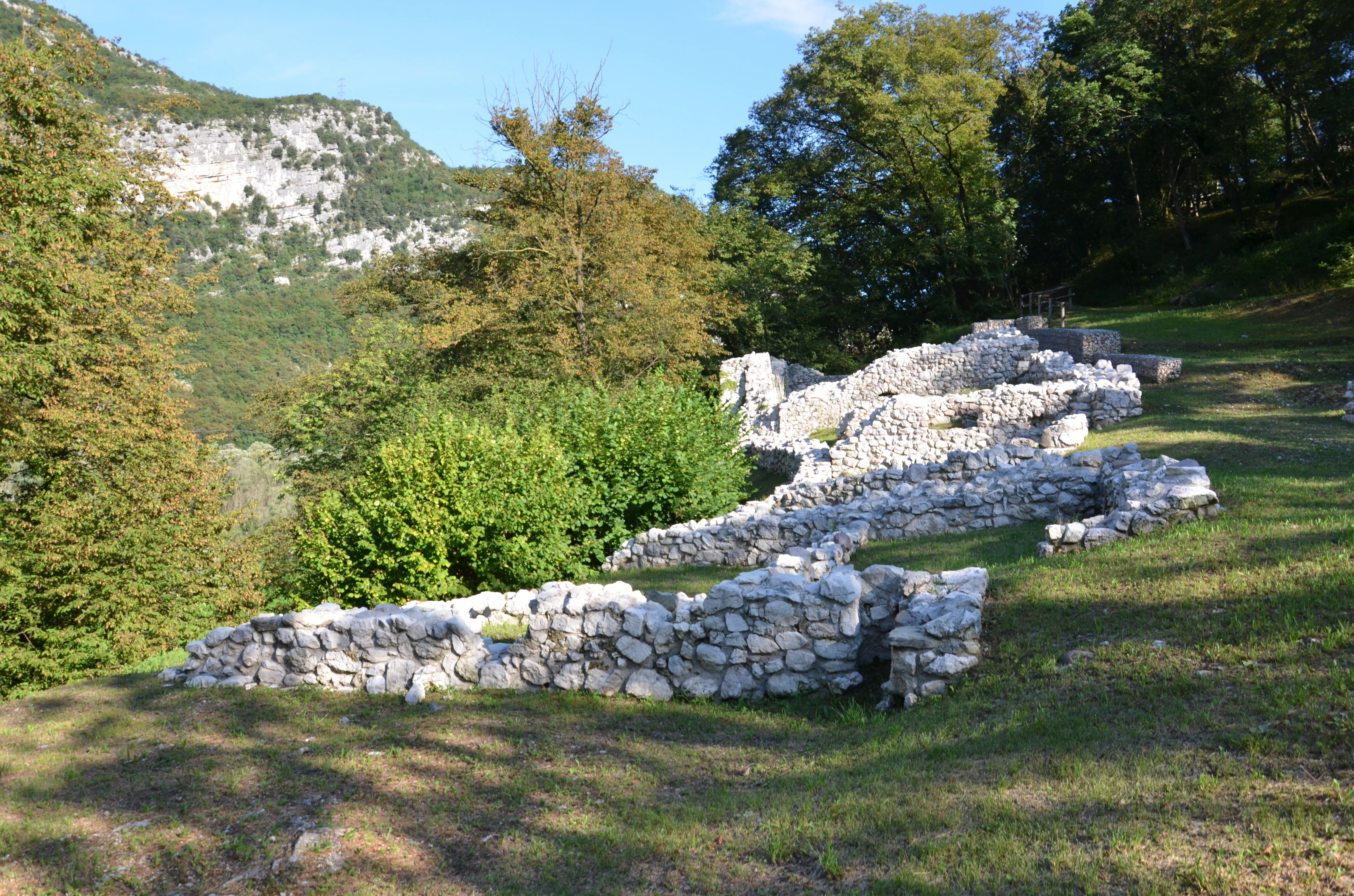 Ancient stone wall ruins located on a grassy hillside, surrounded by trees and mountains at Lago di Loppio.