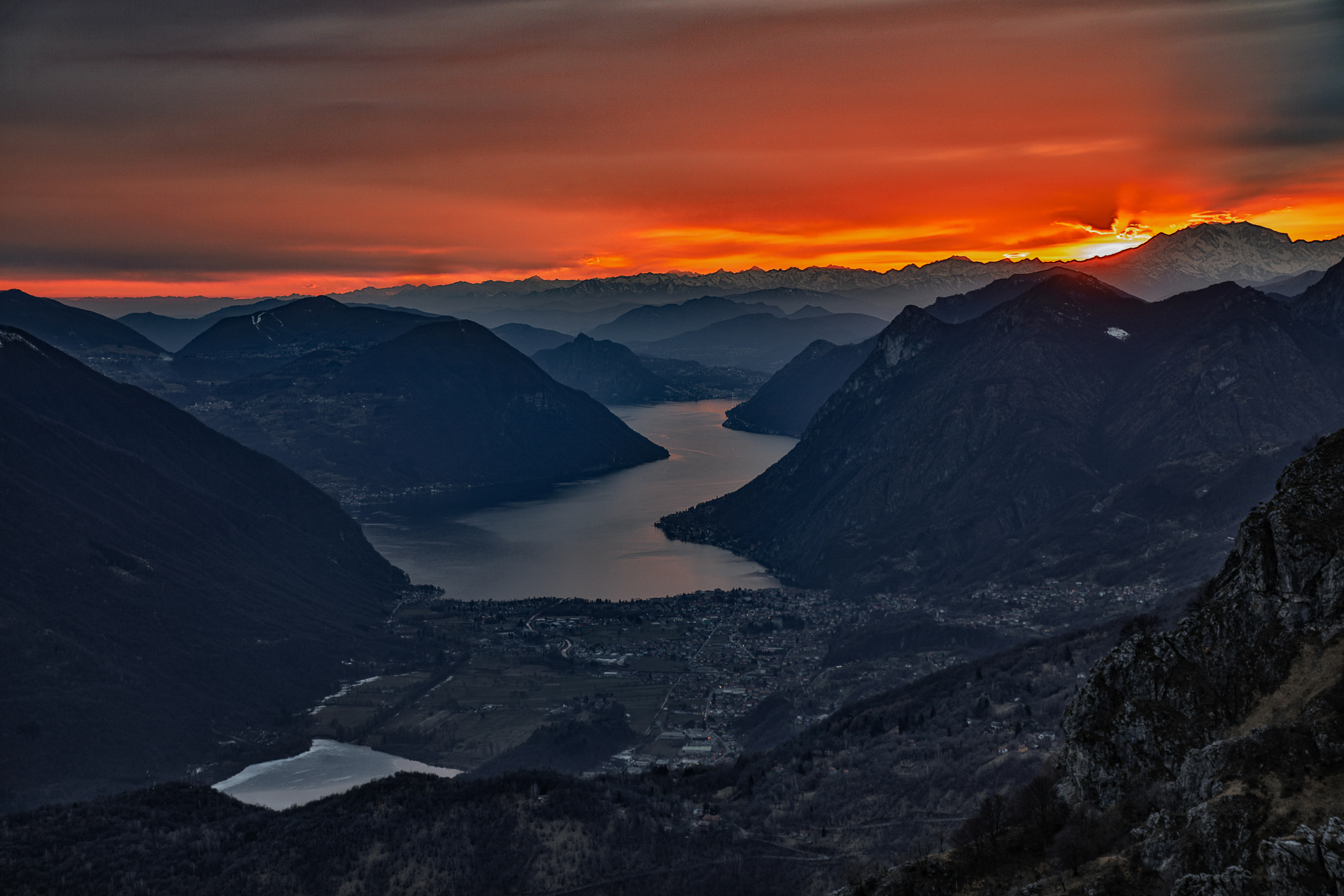 A wide, high-angle landscape shot of a narrow lake nestled between steep, dark mountain ridges during a vibrant sunset with orange and red clouds.