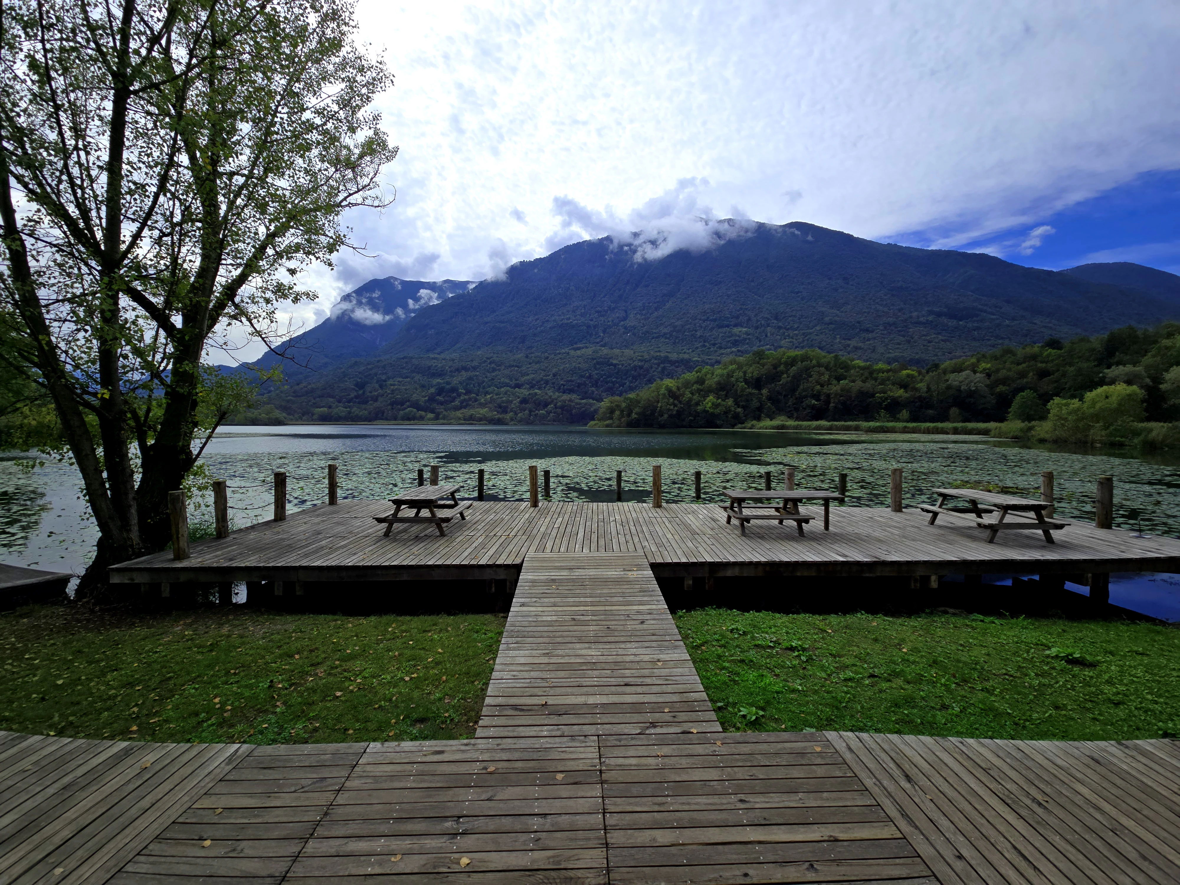 A wide angle view of a wooden observation deck extending into a lake, featuring several picnic tables and a view of mountains under a cloudy sky.