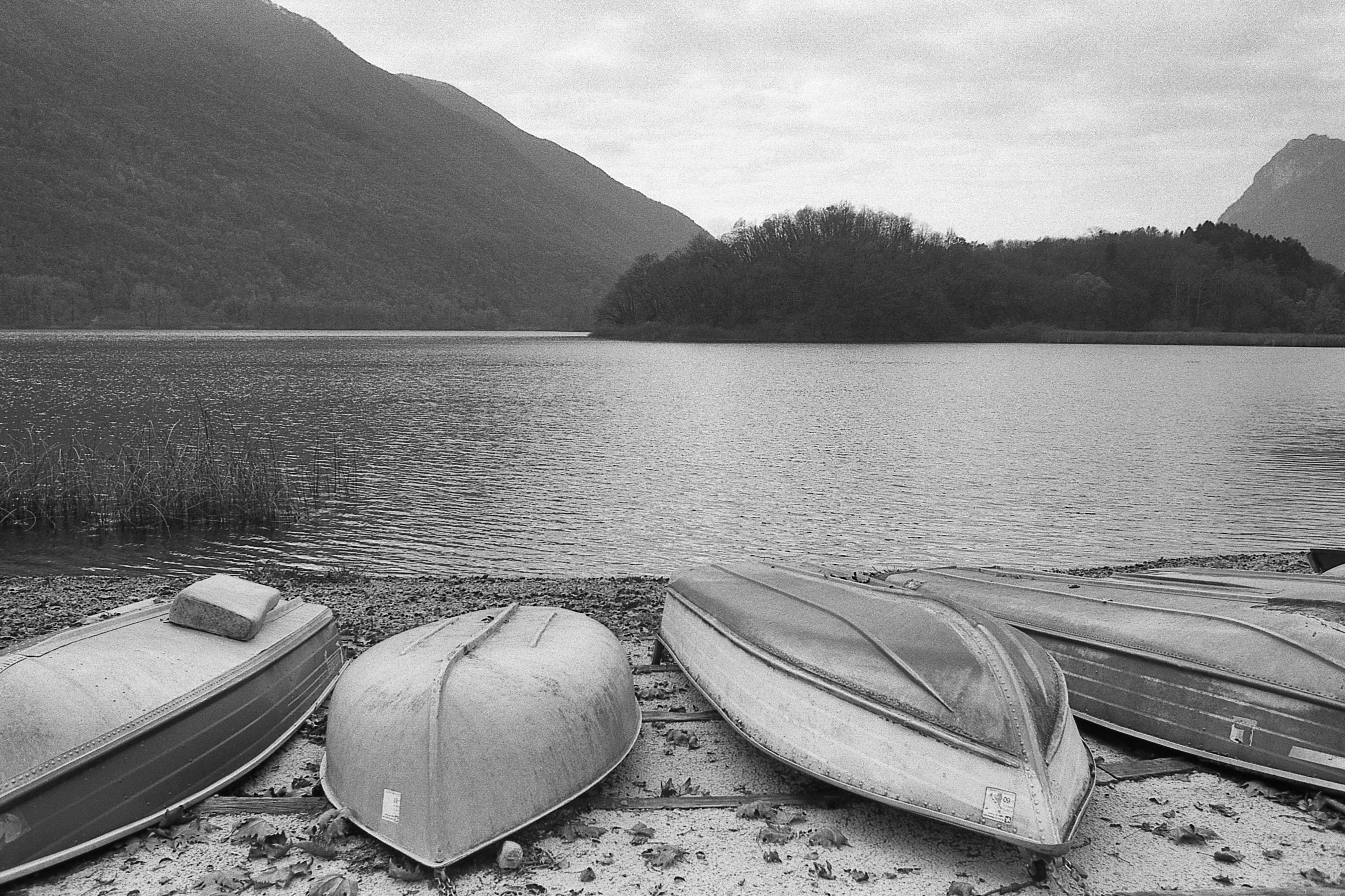 A black and white landscape showing several upturned rowboats on a pebbly lakeshore, with a calm lake and rolling, tree-covered hills in the background.