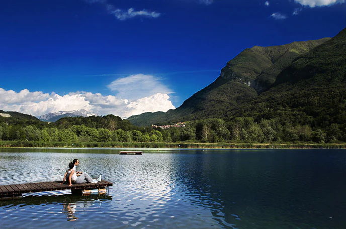 A person sits on a wooden dock extending into a calm blue lake, with dense green trees and large mountain peaks under a clear blue sky with white clouds.