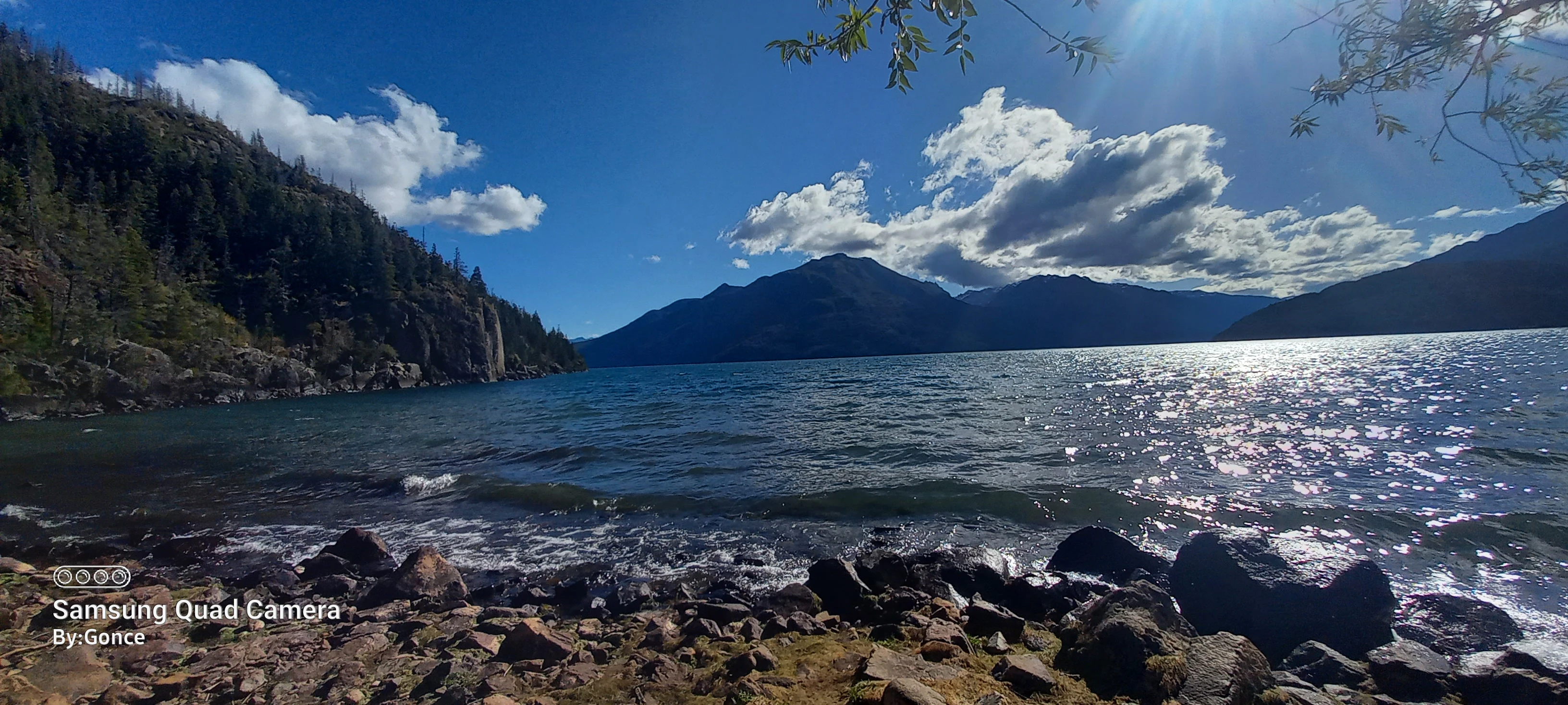 Lake with rocky shore, mountain range in background, blue sky with scattered clouds, sunlight reflecting on water.