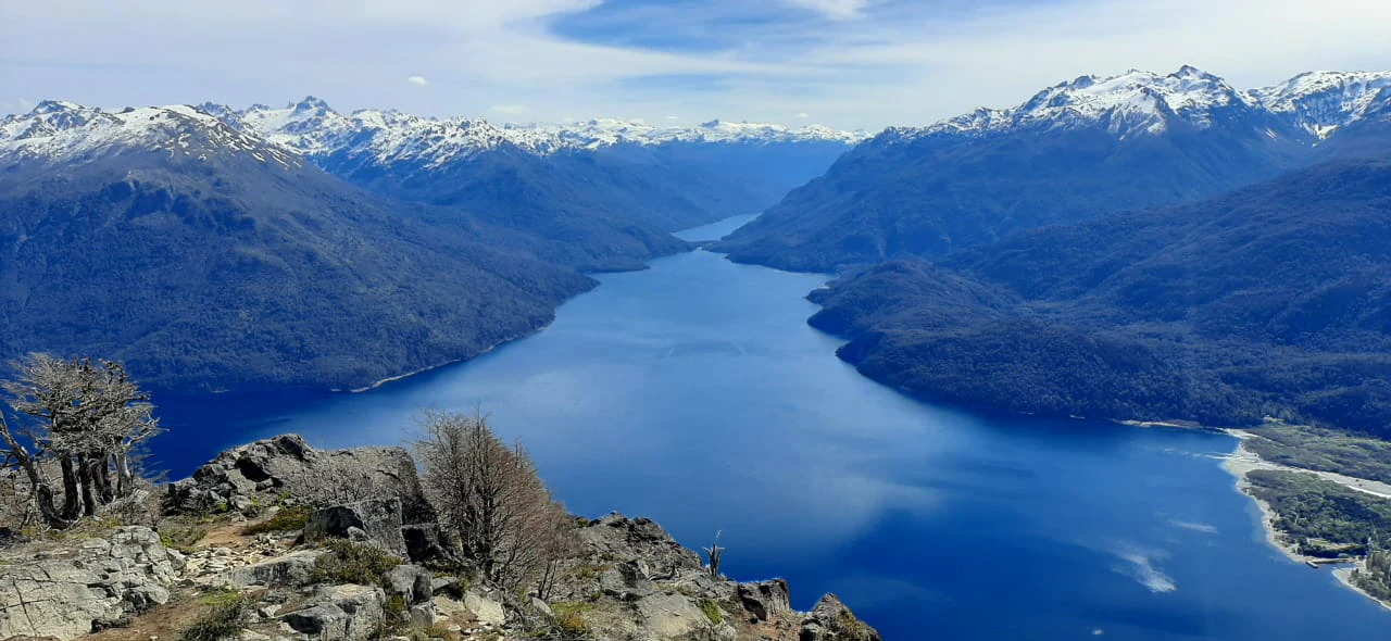 Wide view of a large blue lake surrounded by snow-capped mountains with rocky terrain and sparse trees in the foreground