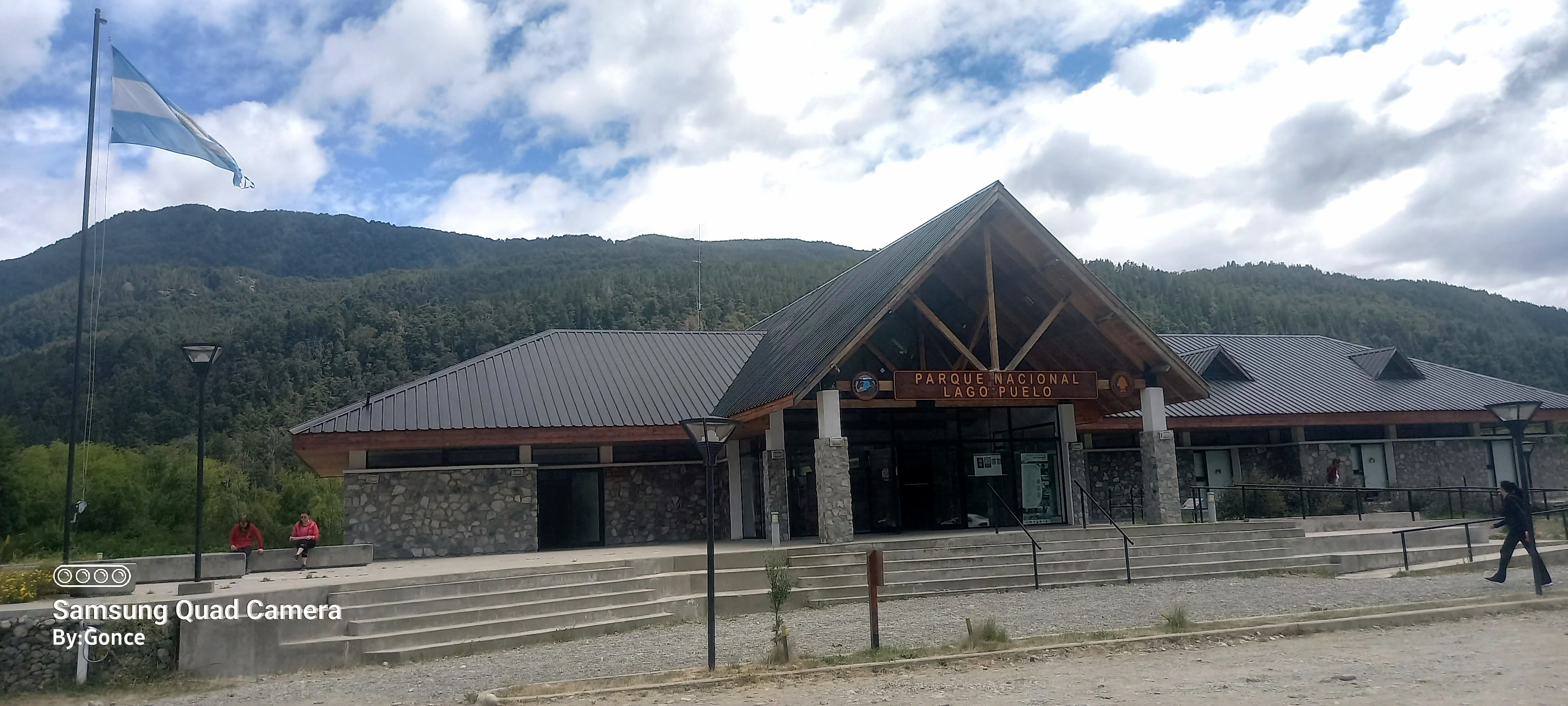 Stone and wood interpretive center building at park entrance with steps, flagpole, mountains, and visitors