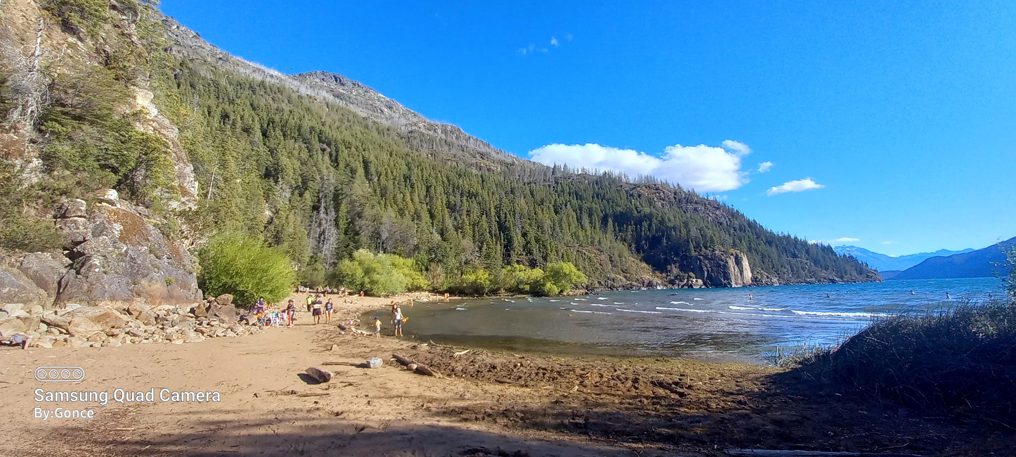 Sandy beach area with forested hills and lake water under clear blue sky