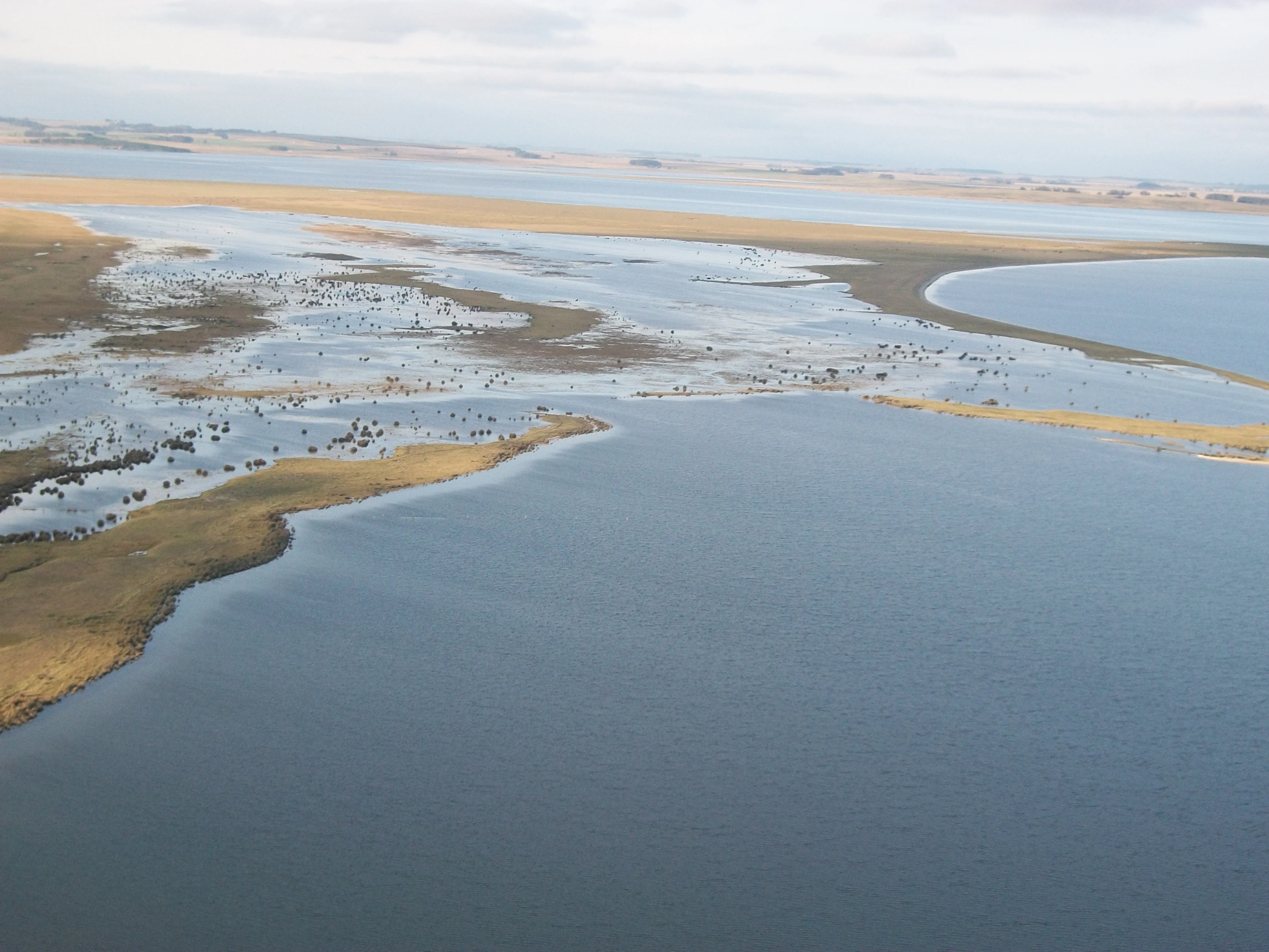 An aerial view of a coastal lagoon with marshy shorelines, sandy patches, and open water under a soft, overcast sky.