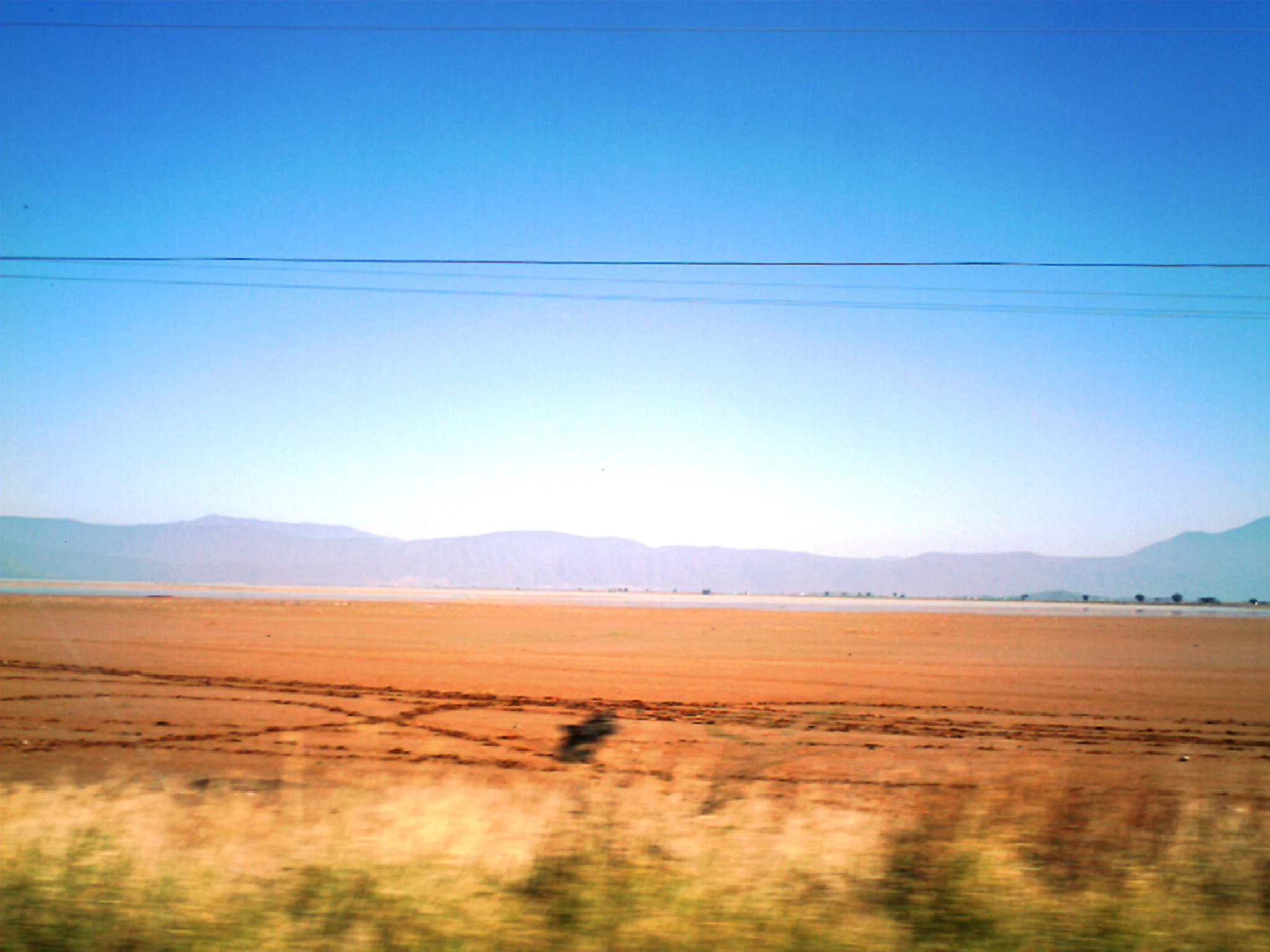 A vast, flat, and arid dry lake bed under a clear blue sky, with rolling mountains in the distance and visible power lines stretching across the top of the frame.