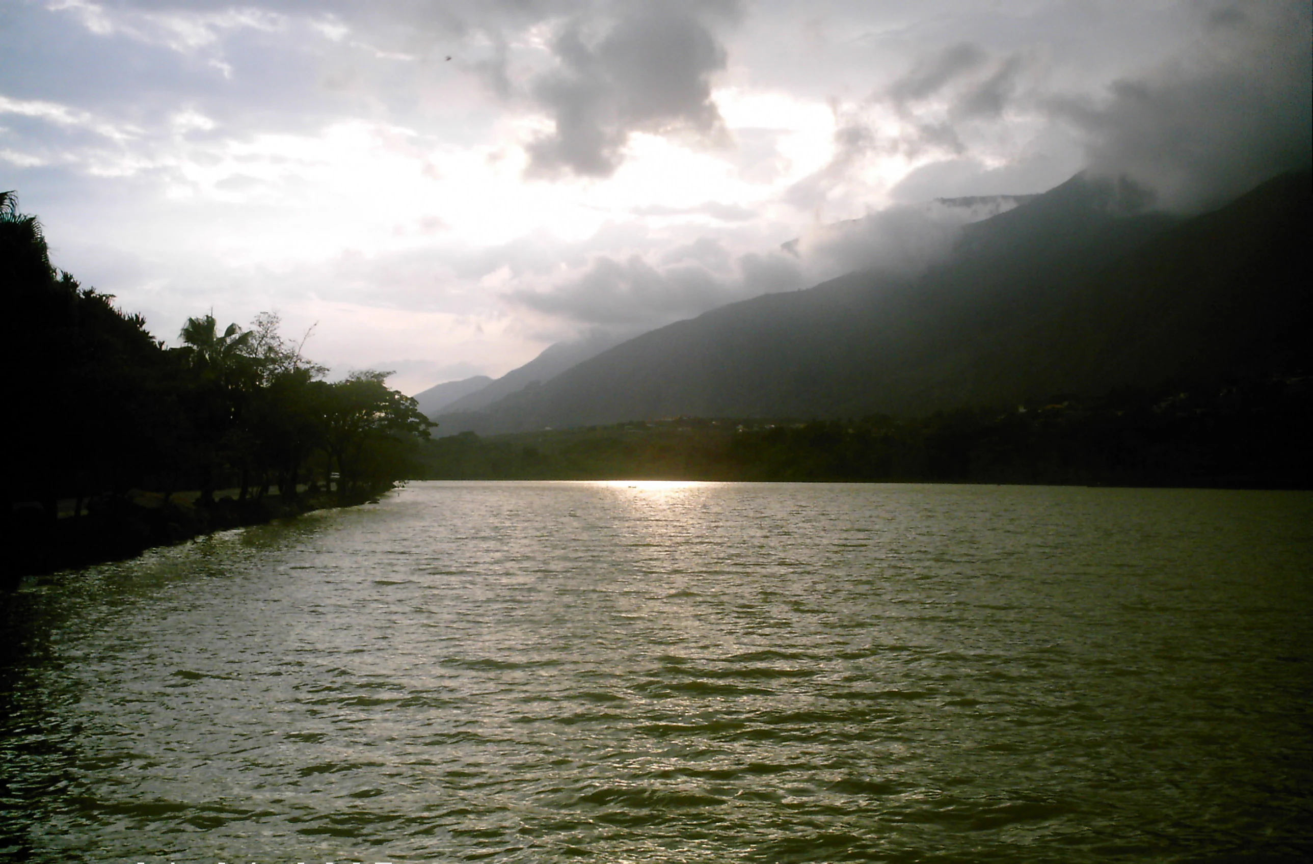 Una vista amplia de un lago que refleja el sol con montañas al fondo bajo un cielo nublado.