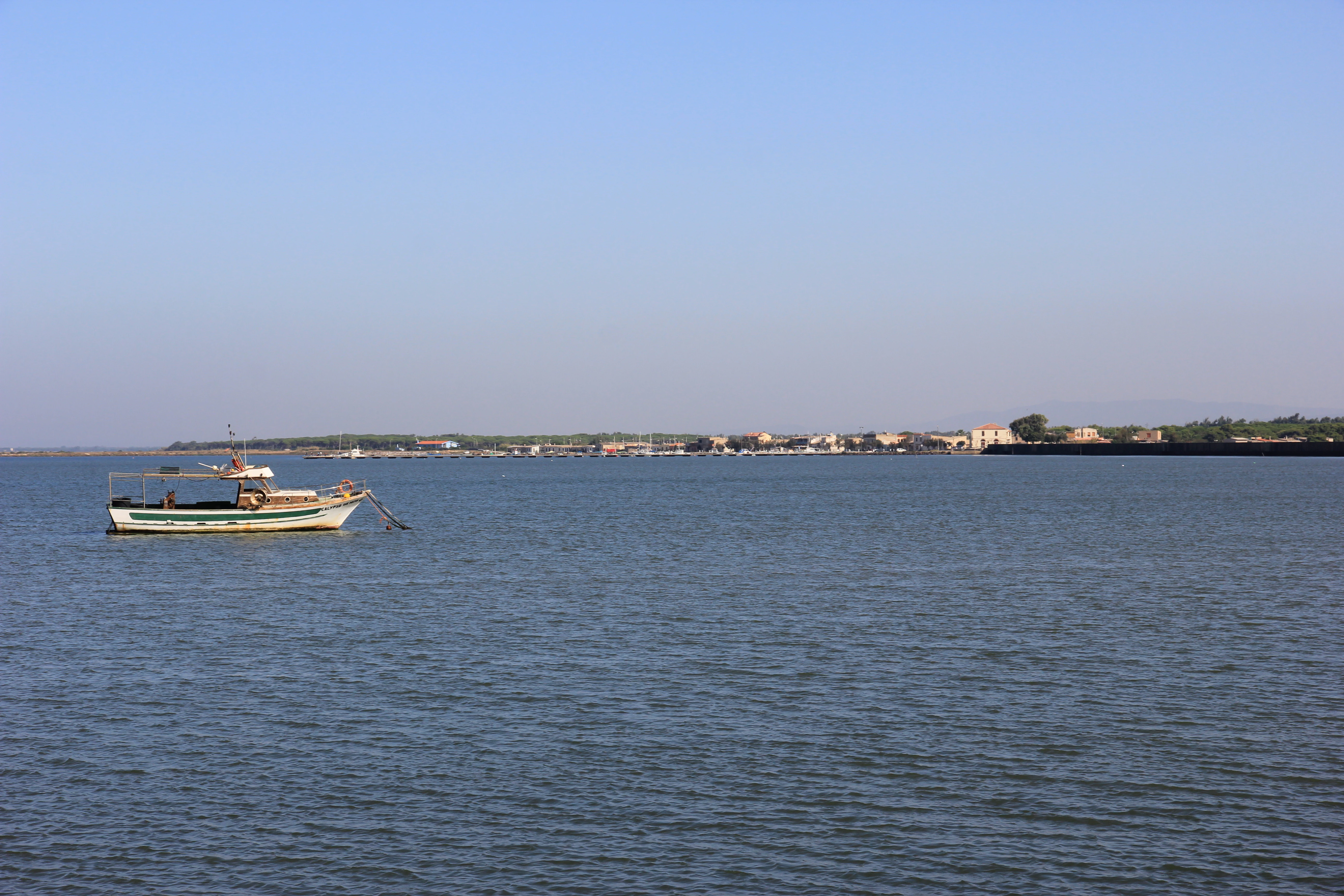 Vista amplia de una laguna tranquila con un pequeño bote de pesca anclado en el primer plano izquierdo y una costa lejana con edificios al fondo bajo un cielo despejado.