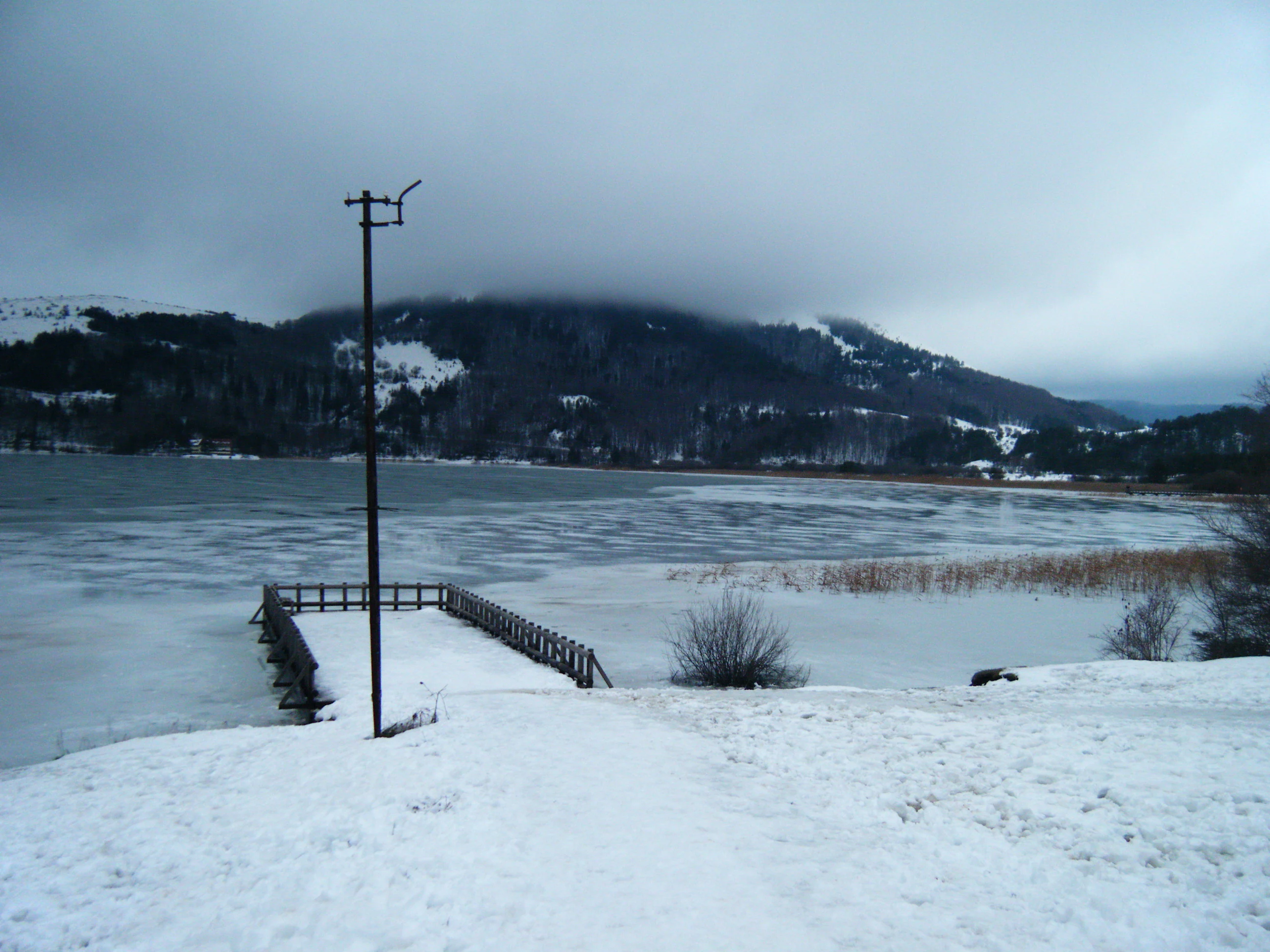 Frozen lake with wooden pier extending into icy water, snow-covered ground, mountains in background under overcast sky