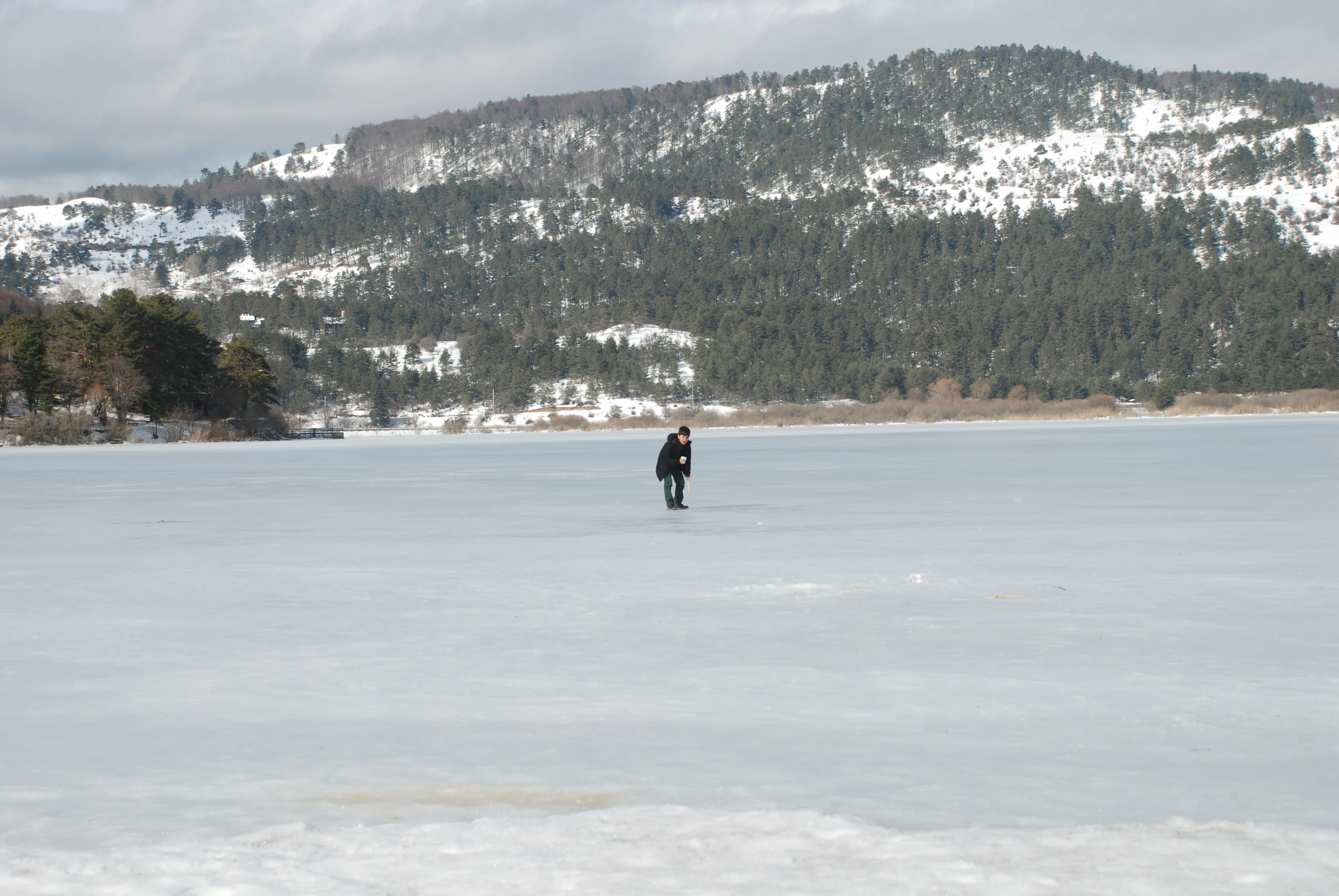 Frozen lake surface with a person standing on ice, snow-covered hills and trees in the distance