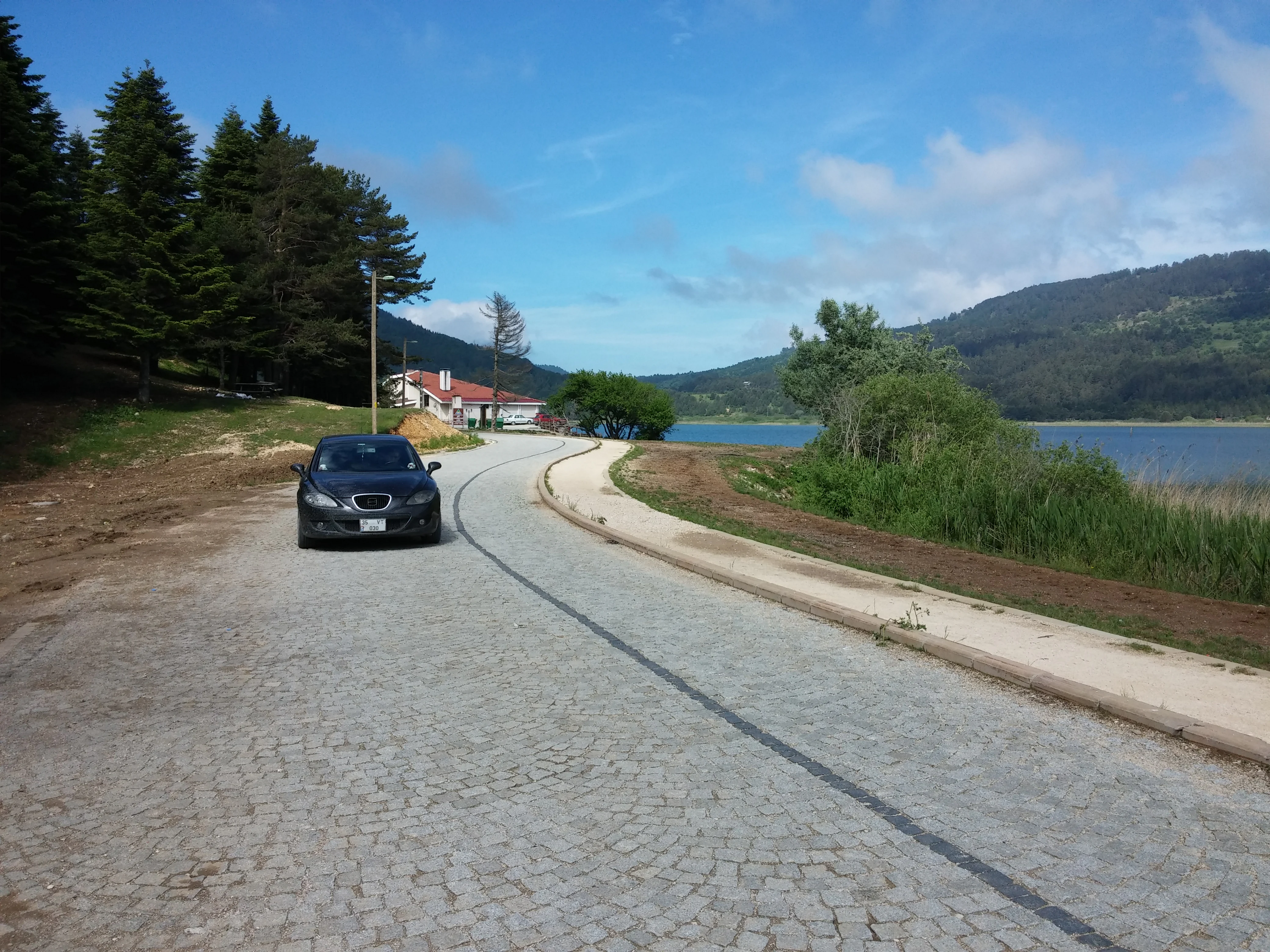 Cobblestone road by a lake with mountains in the distance, trees on the left, and a car on the road