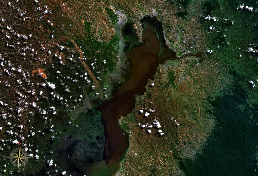 Top-down satellite imagery showing the dark water of Lake Alaotra surrounded by green vegetation and marshland, with scattered white clouds.