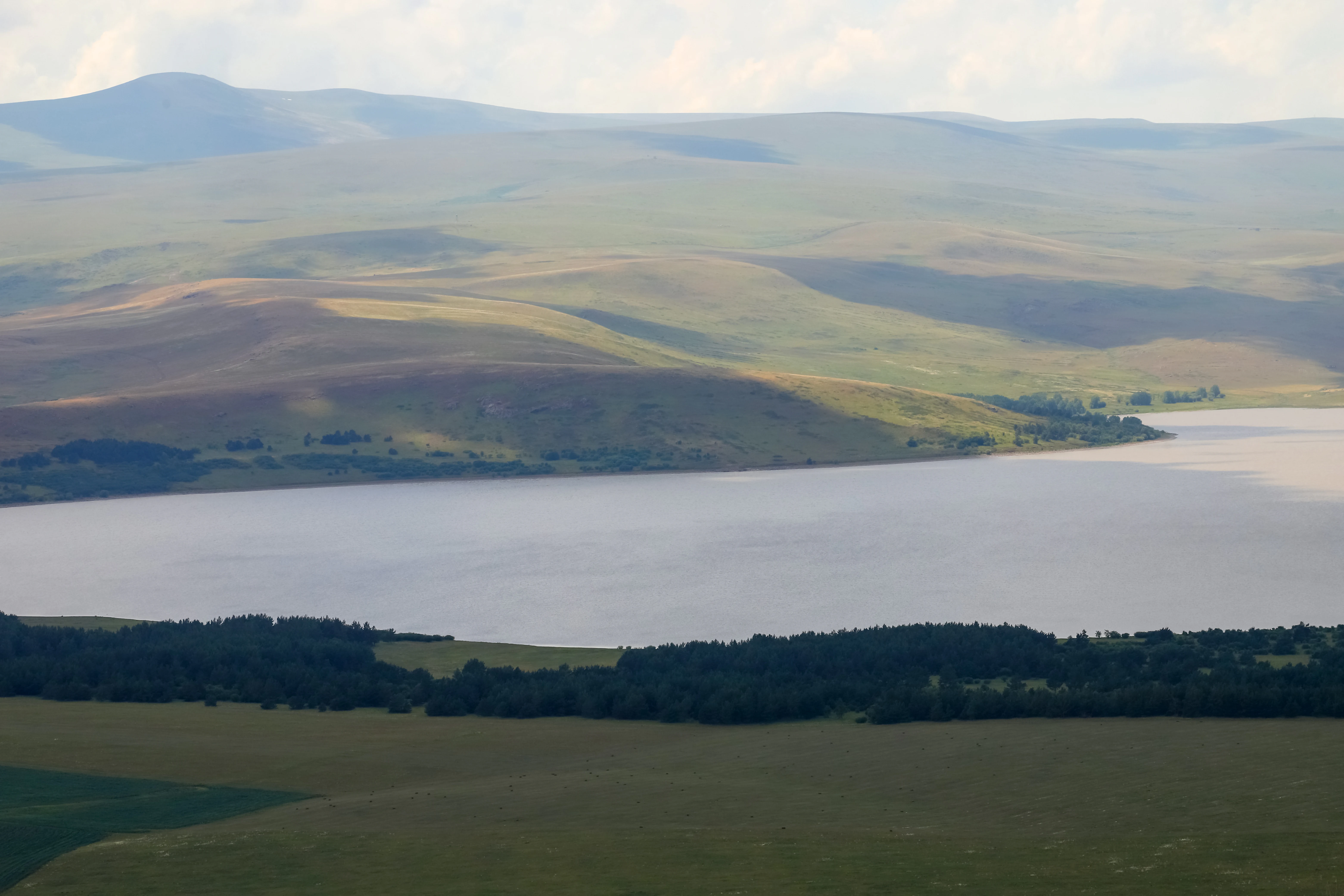 A wide landscape view featuring a large lake surrounded by rolling hills and green fields under a partly cloudy sky
