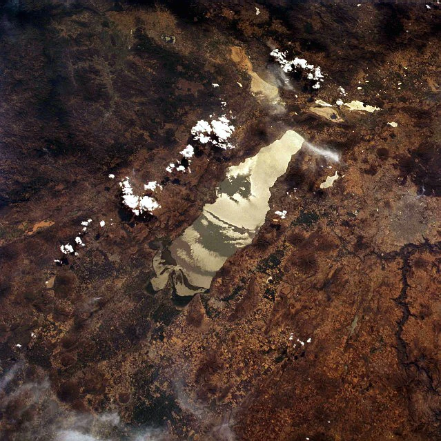 Satellite view of Lake Chapala, Mexico, surrounded by brown terrain with scattered white clouds floating above.