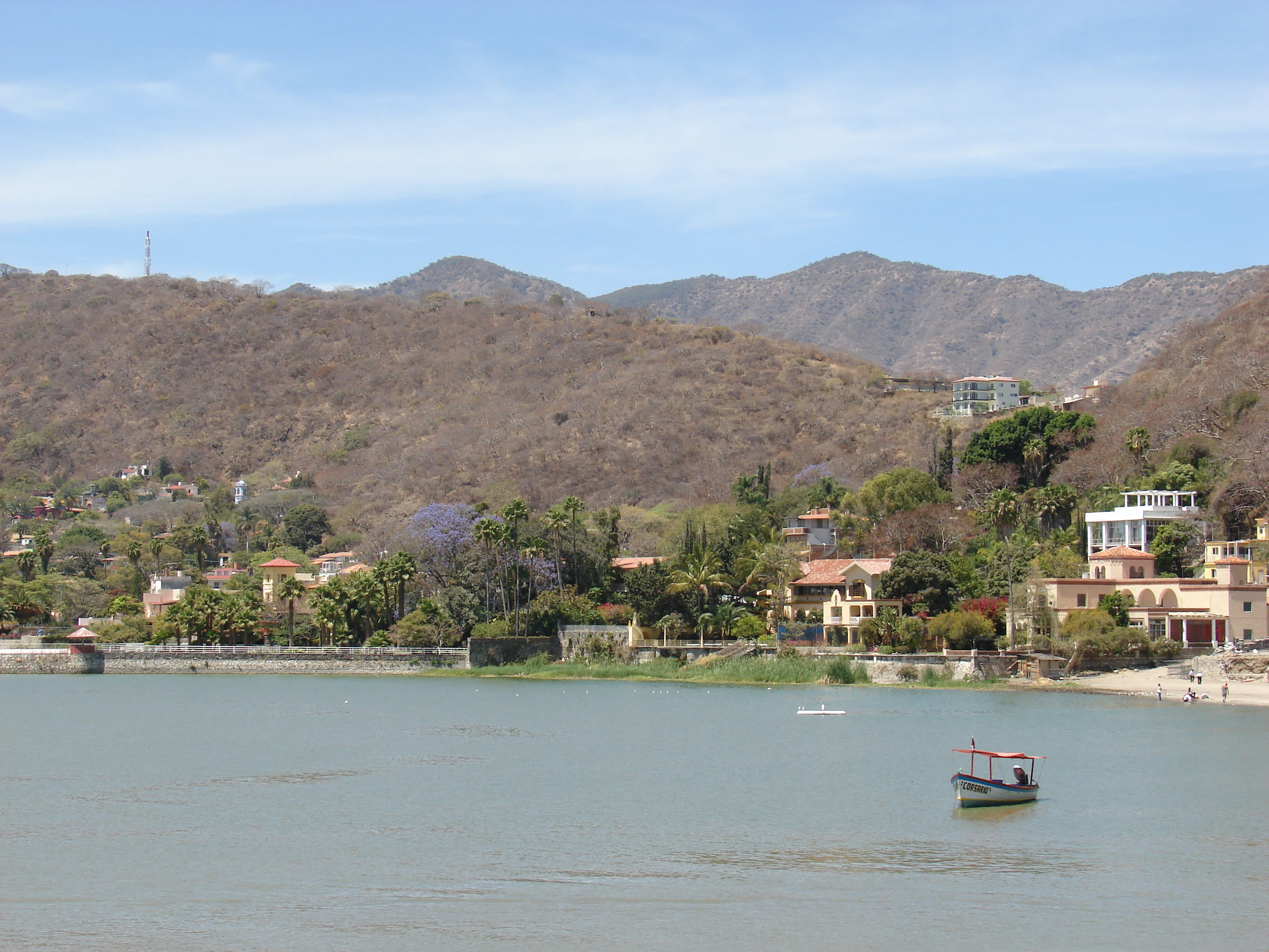 A wide view of Lake Chapala with a small boat in the foreground and a hillside town with houses and dry vegetation in the background.