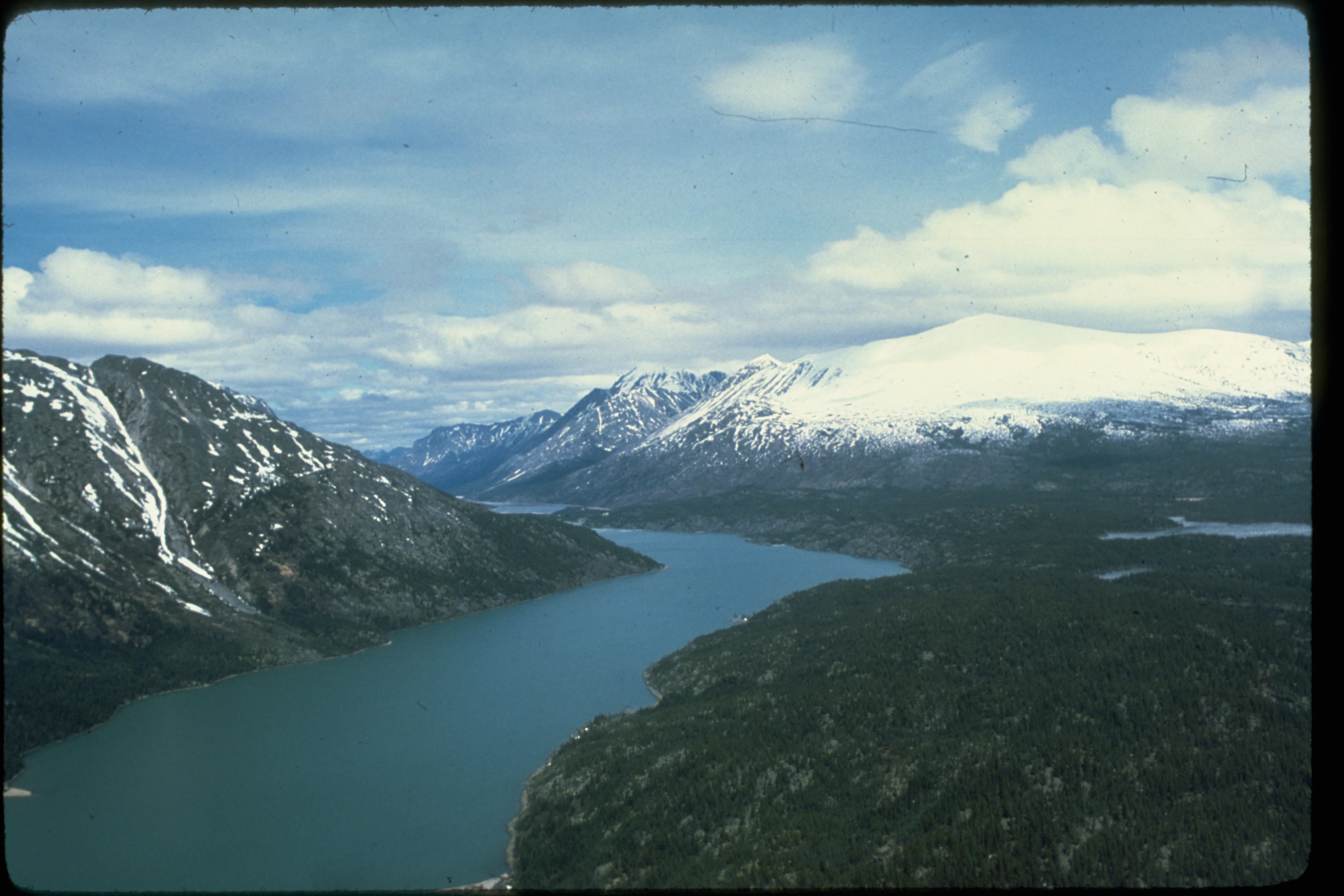 An aerial shot of a long, narrow lake winding through a valley, flanked by tall, snow-capped mountains and dense coniferous forest.